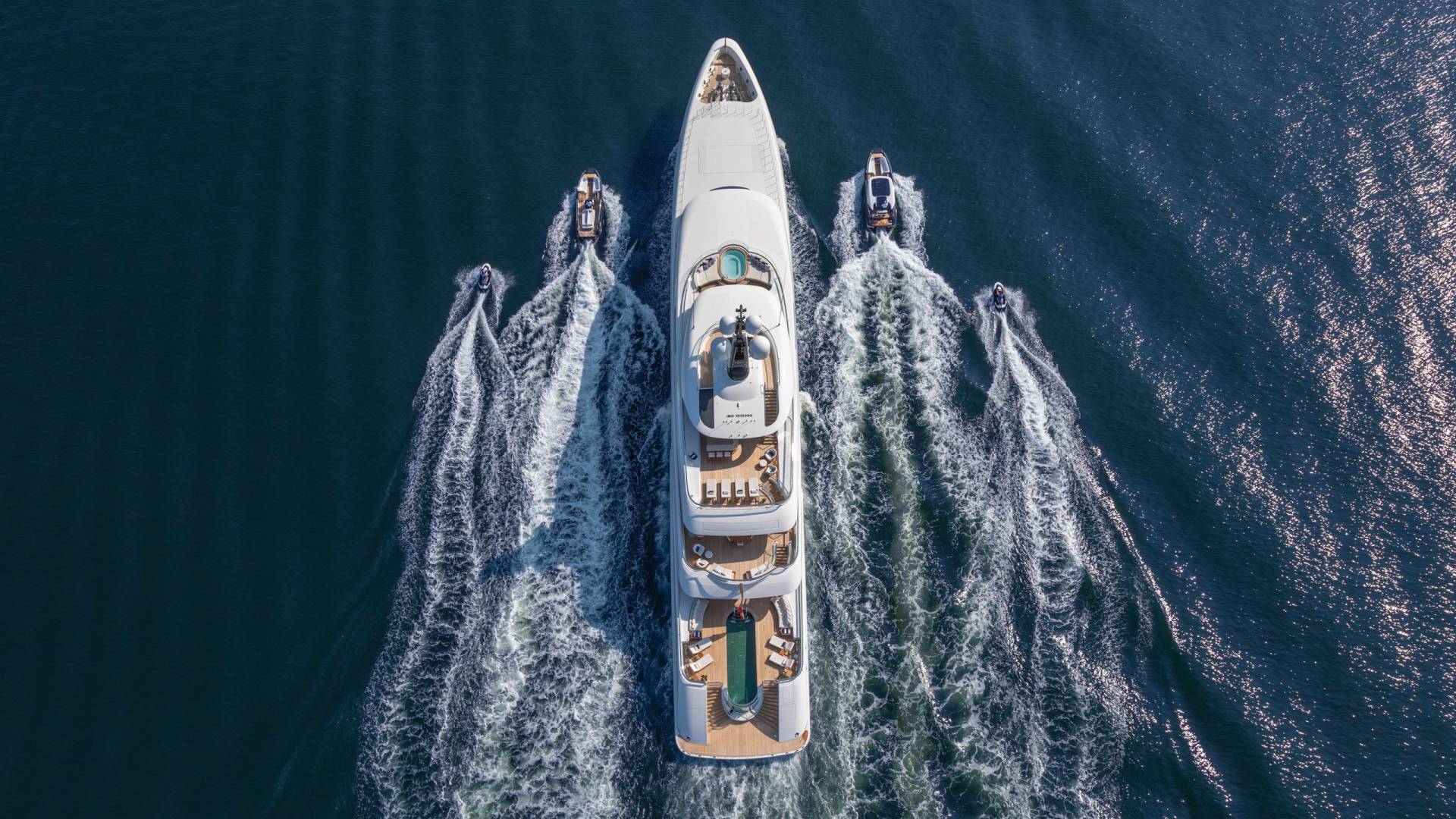 A luxurious superyacht cruises through the deep blue ocean, trailed by smaller vessels creating white wakes.