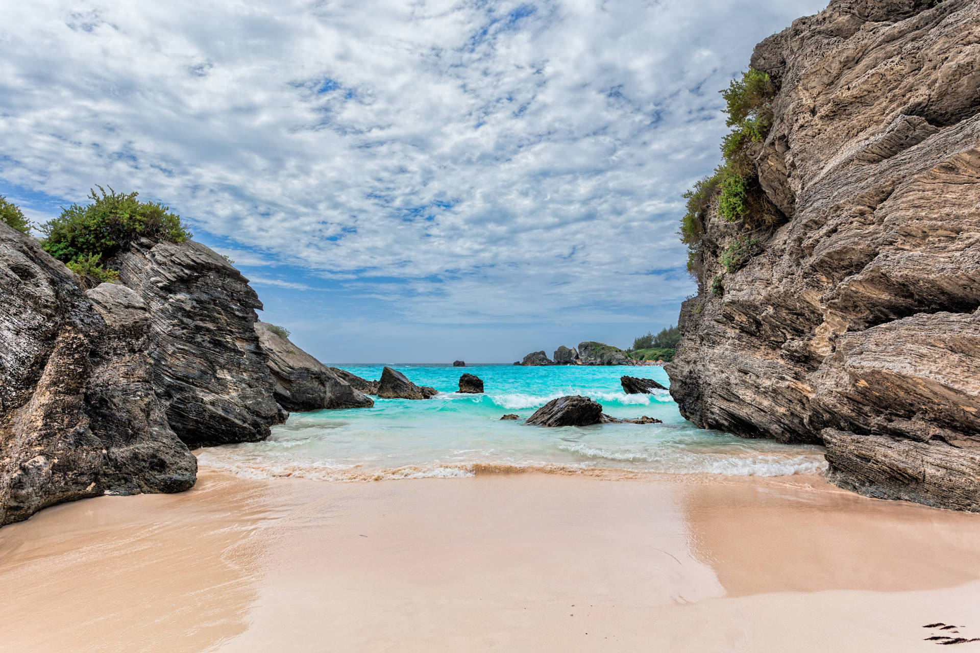 Turquoise waters gently lap onto a pink sand beach framed by dramatic rock formations under a cloudy sky.
