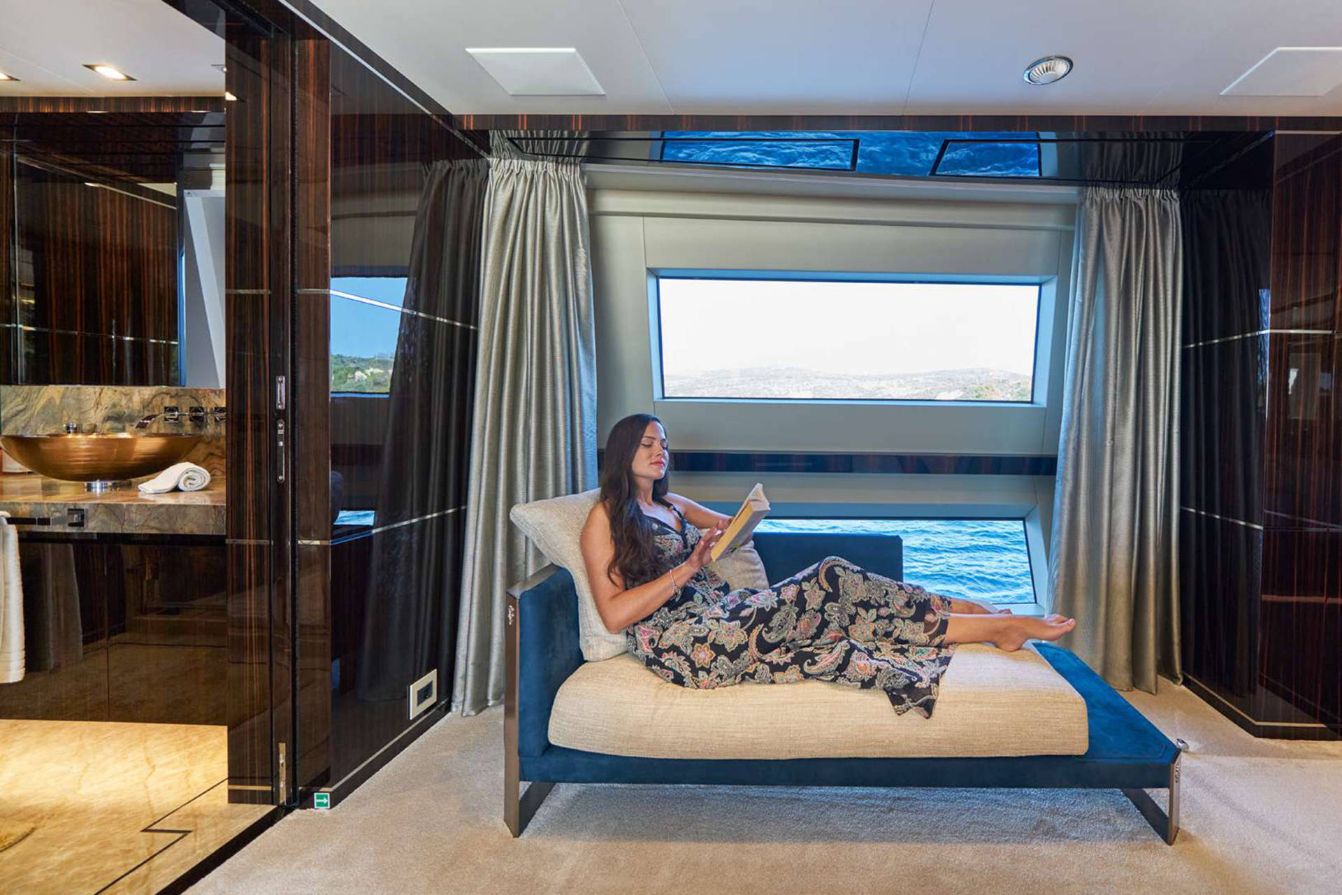 A woman relaxes with a book on a chaise lounge aboard a luxurious yacht, with the sea visible through a panoramic window.