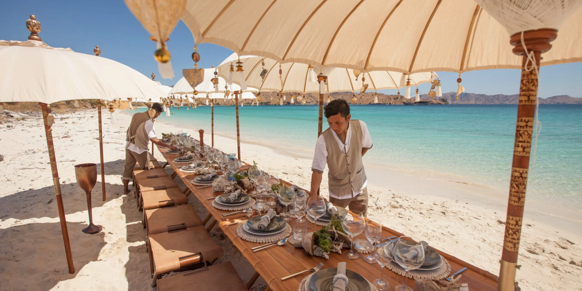 A long, beautifully set table awaits guests on a pristine beach, shaded by ornate umbrellas and overlooking turquoise waters.