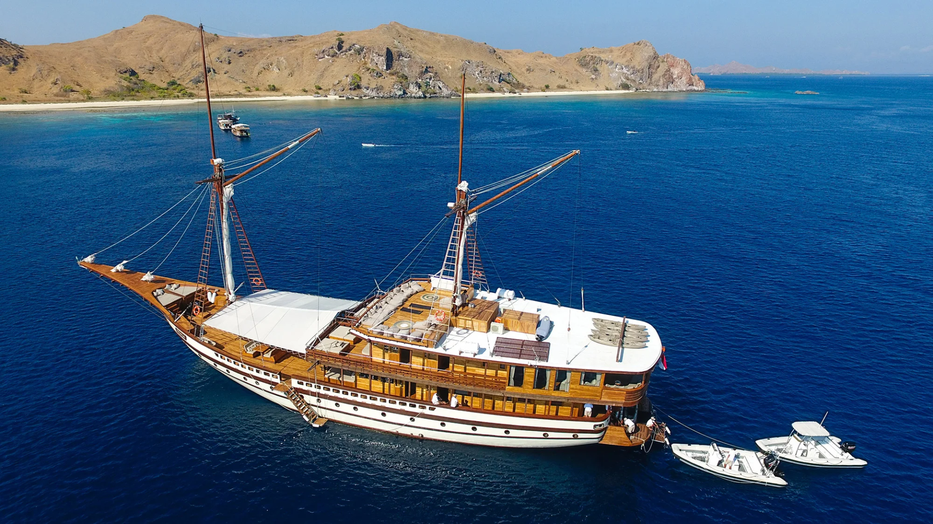 A majestic wooden schooner sails on a tranquil blue sea, anchored near a rugged, arid coastline.