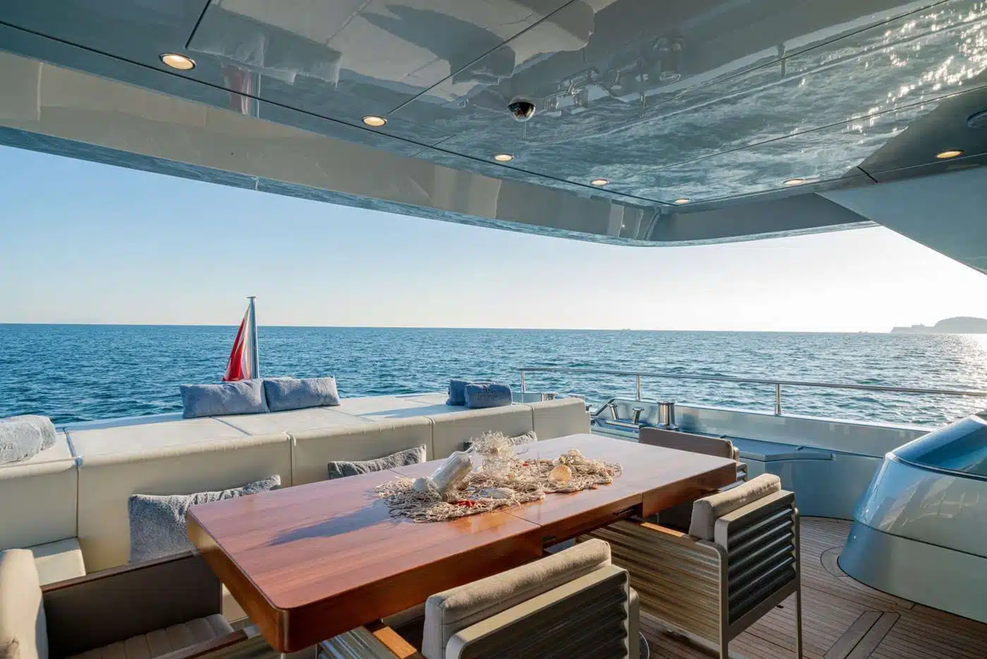 A luxurious yacht deck with a wooden table and chairs, overlooking the serene blue ocean and clear sky in the background.