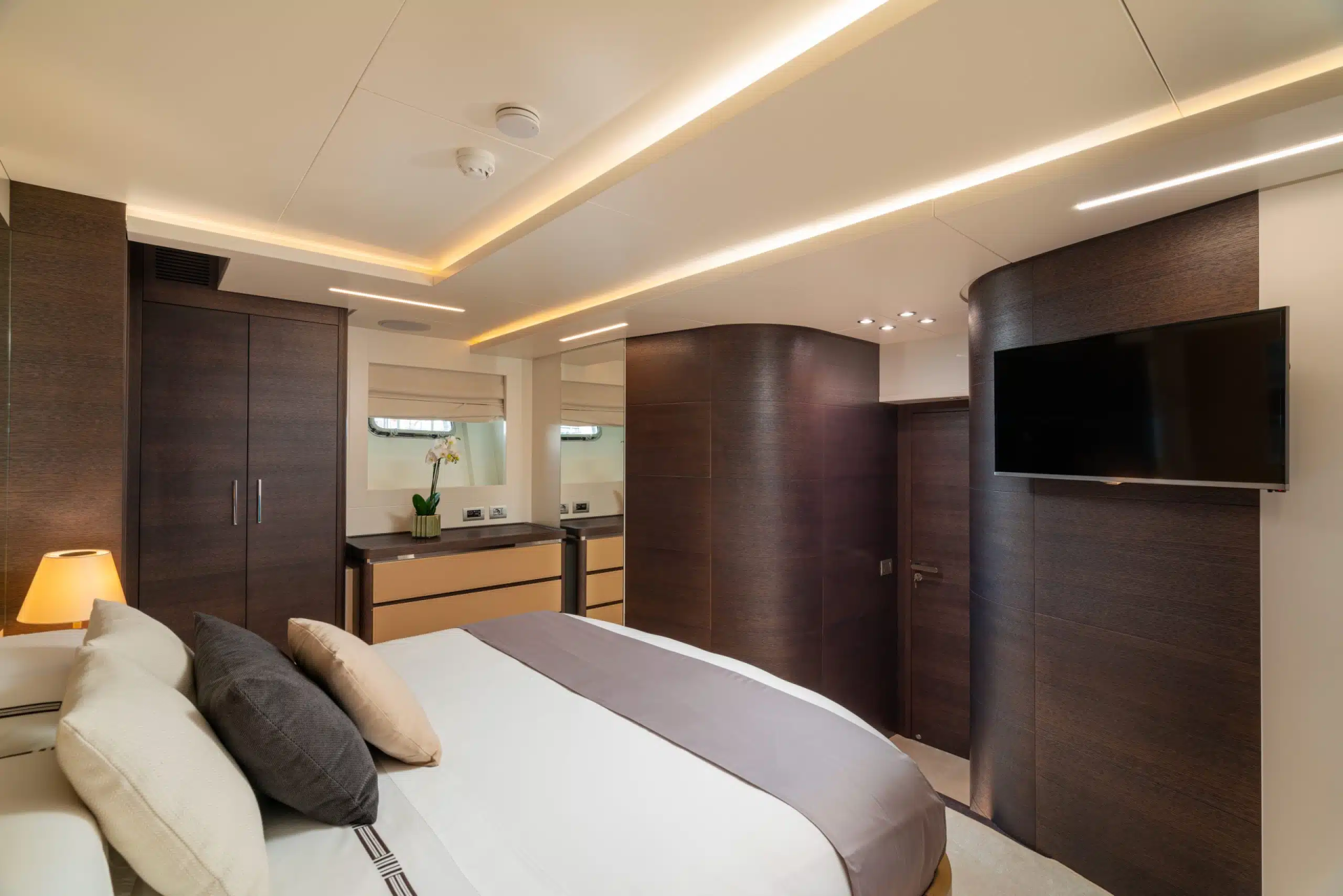 A modern and luxurious bedroom with a large bed, dark wood furniture, and a television mounted on the wall. The room features recessed lighting and a minimalist design aesthetic.