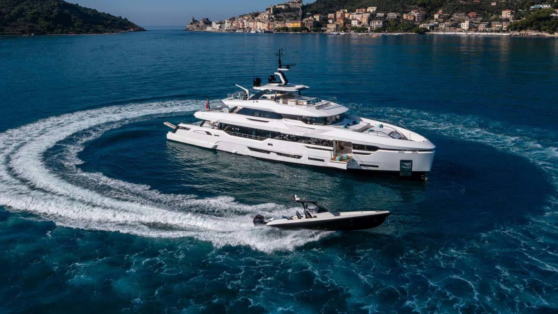 A large white yacht cruises through the deep blue sea, leaving a swirling wake, while a smaller boat speeds ahead.