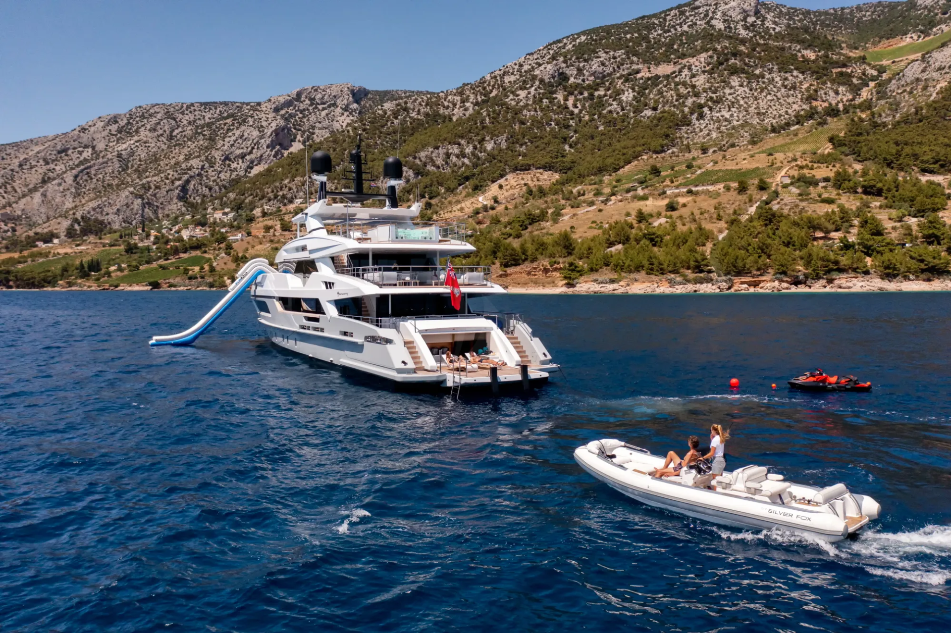 A luxurious yacht with a water slide is anchored offshore near a verdant, mountainous coastline.