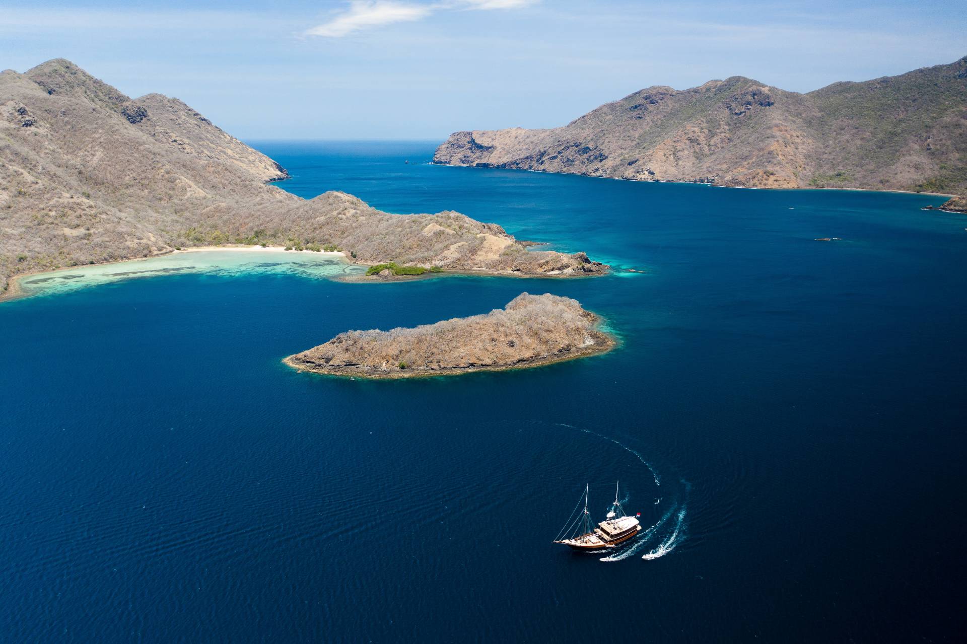 Stunning aerial view of a remote tropical island with rugged mountains, crystal-clear turquoise waters, and a small sailboat anchored in the foreground.