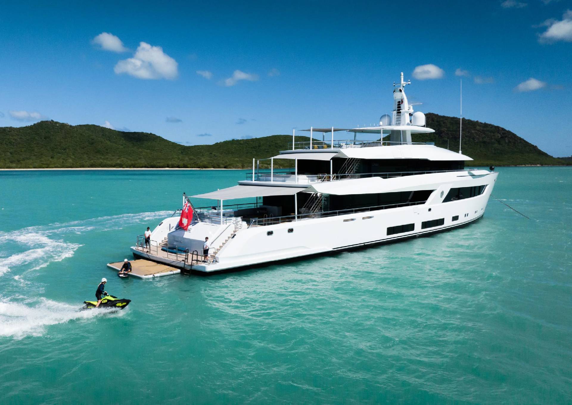 A luxurious white yacht anchors in a tranquil turquoise bay, with a jet ski zipping by and a lush green island in the background.