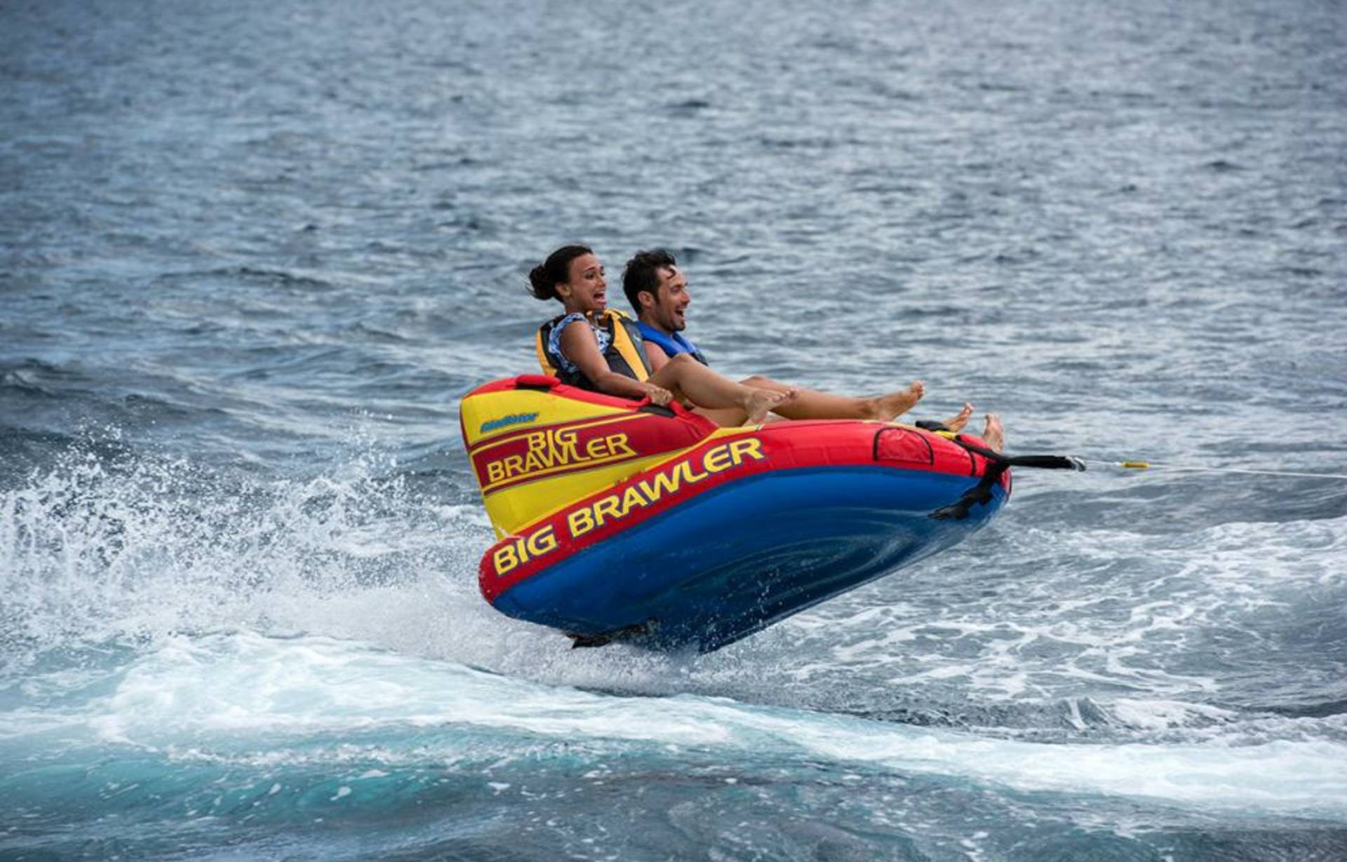 A couple enjoys a thrilling ride on a "Big Brawler" inflatable tube being pulled across the choppy ocean waters.