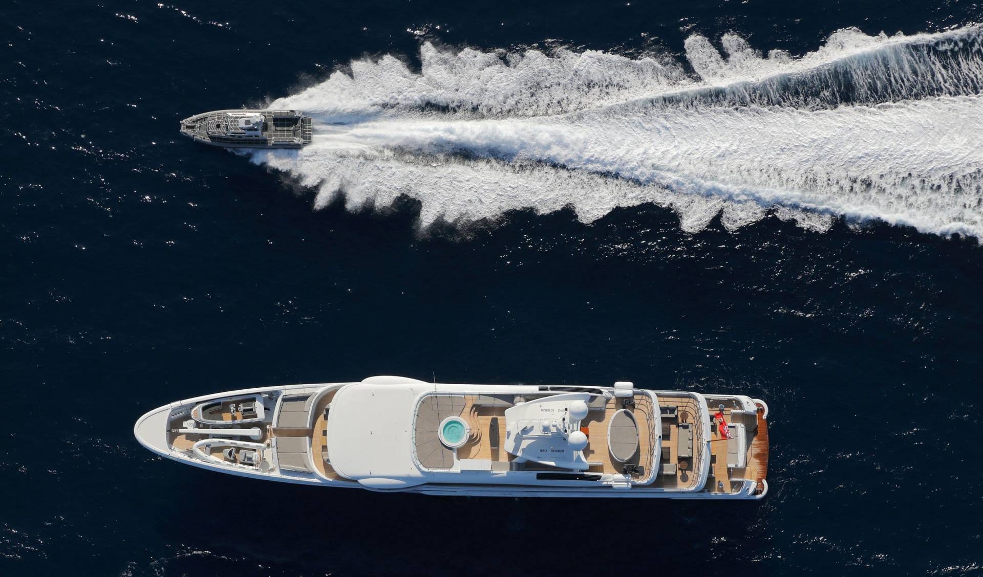 A sleek superyacht glides through the deep blue ocean, leaving a trail of white wake, while a smaller vessel speeds ahead.