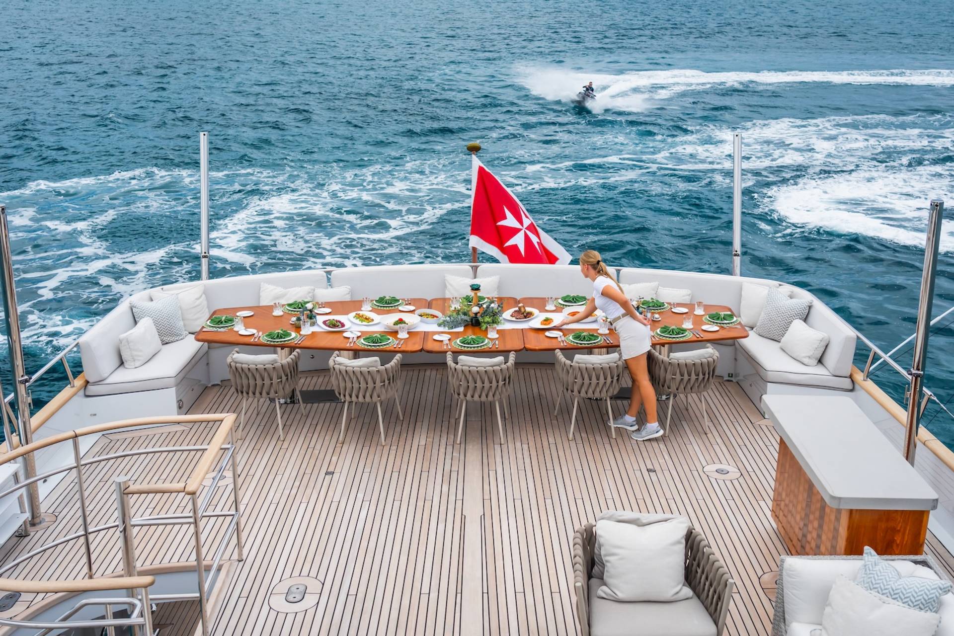 A woman sets a beautifully arranged dining table on a yacht's deck as a jet ski speeds by in the ocean.
