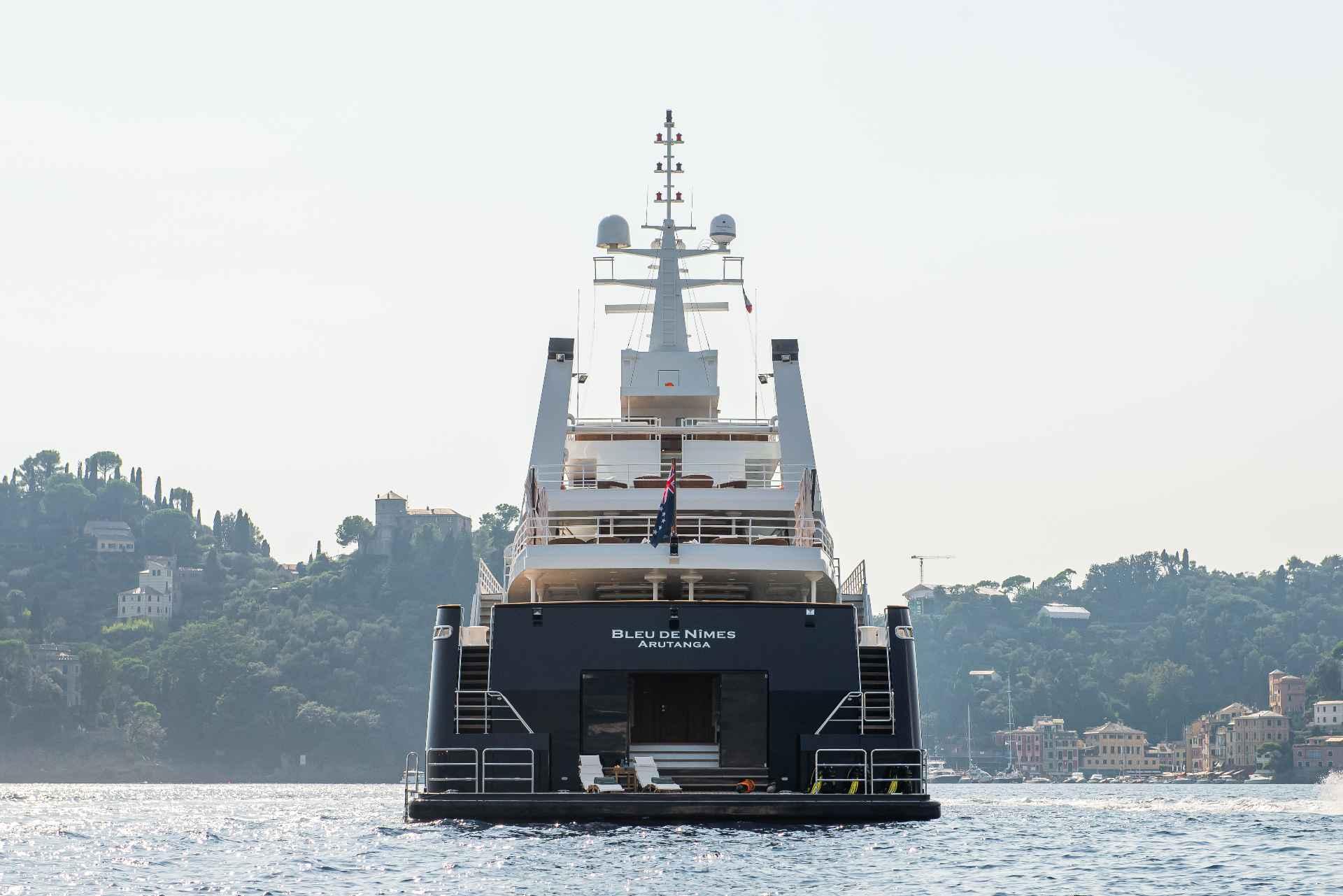 The impressive yacht Bleu de Nîmes cruises past a picturesque coastal town nestled on a lush green hillside.