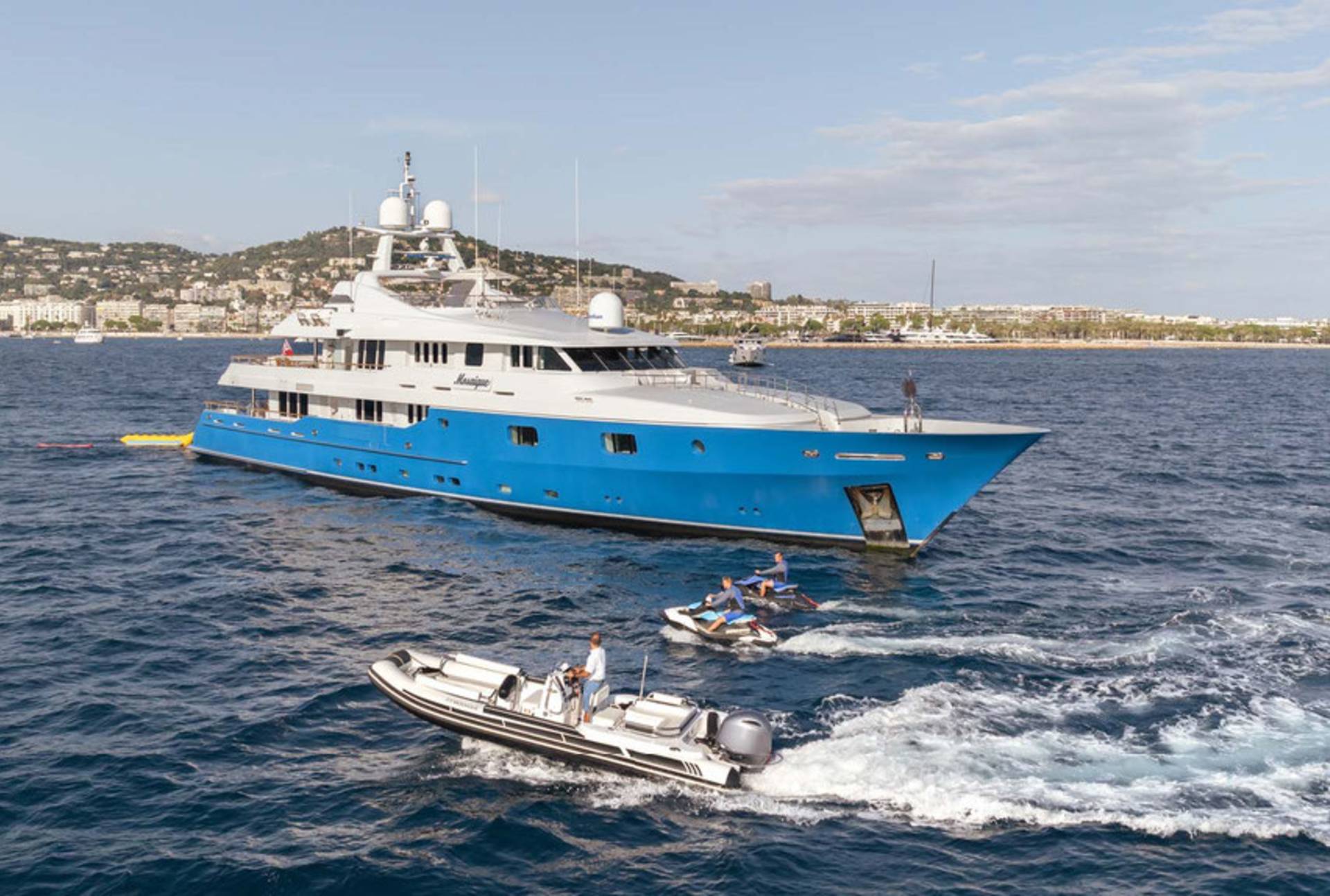 A vibrant blue yacht cruises through the water, accompanied by jet skis and a smaller boat near a coastal city.