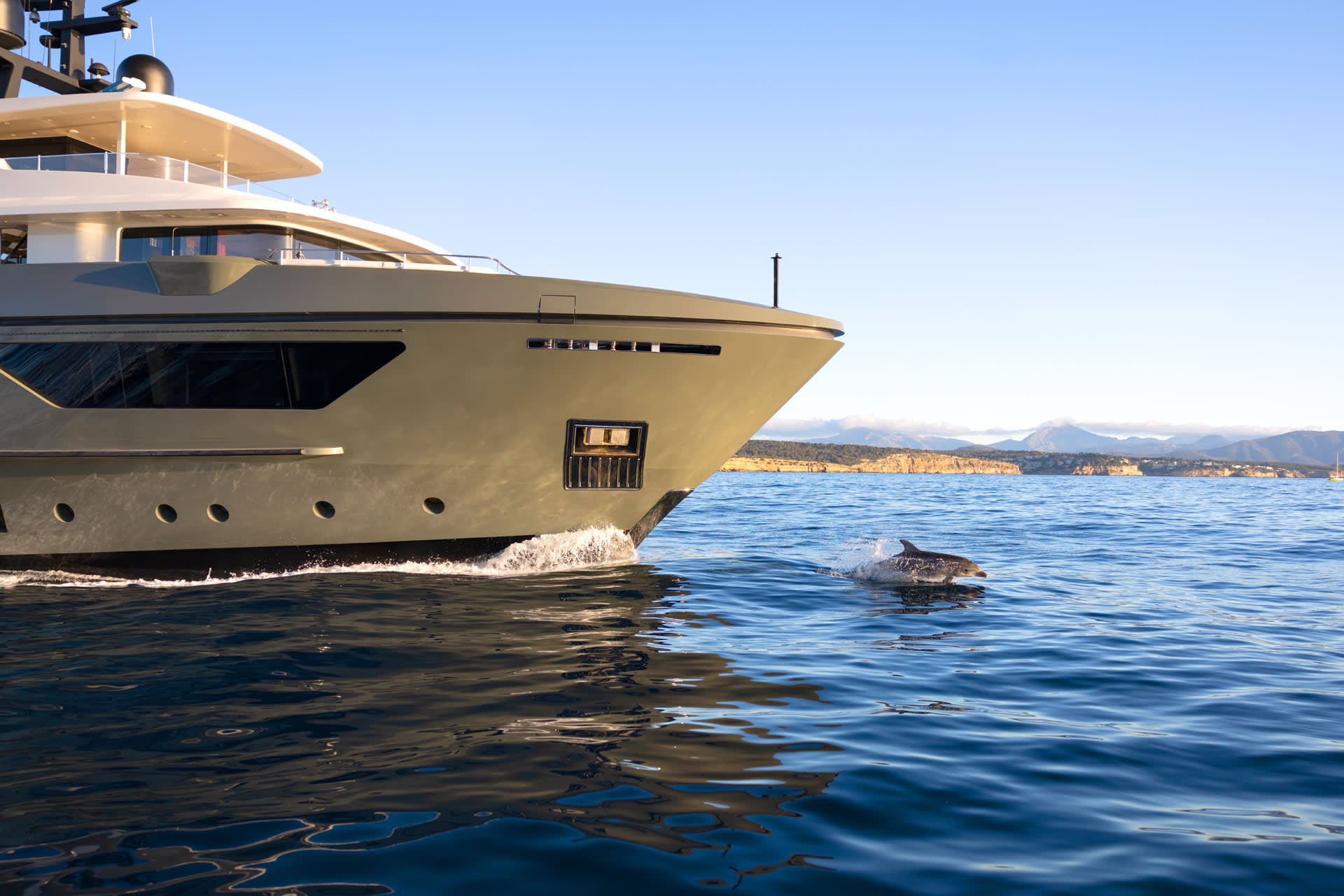 A sleek yacht glides through the calm sea as a playful dolphin leaps from the water beside it.