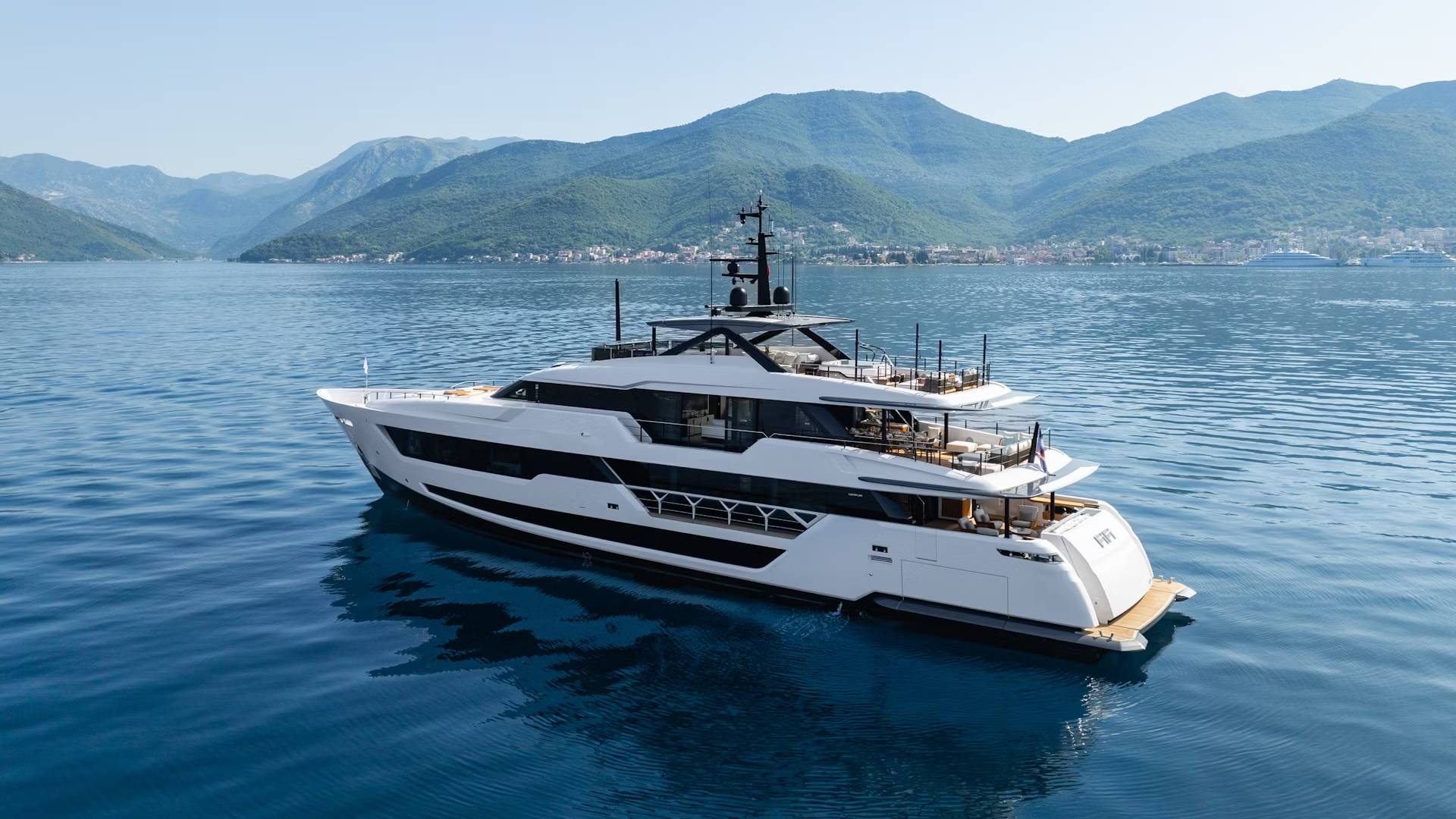A sleek white superyacht glides through calm blue waters with lush green mountains in the background.