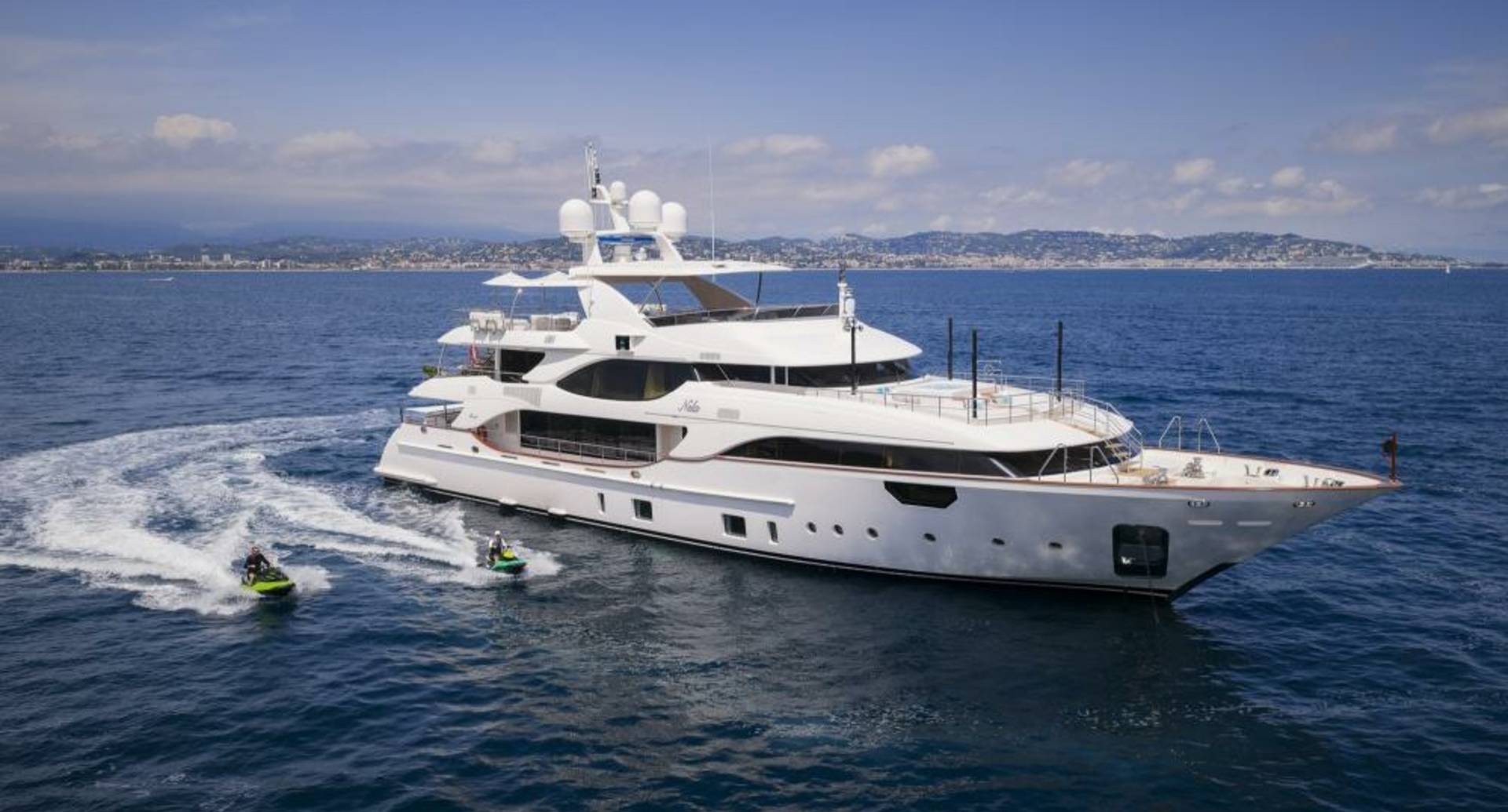 A luxurious white yacht cruises through the azure waters with jet skiers trailing behind, a picturesque coastline in the distance.