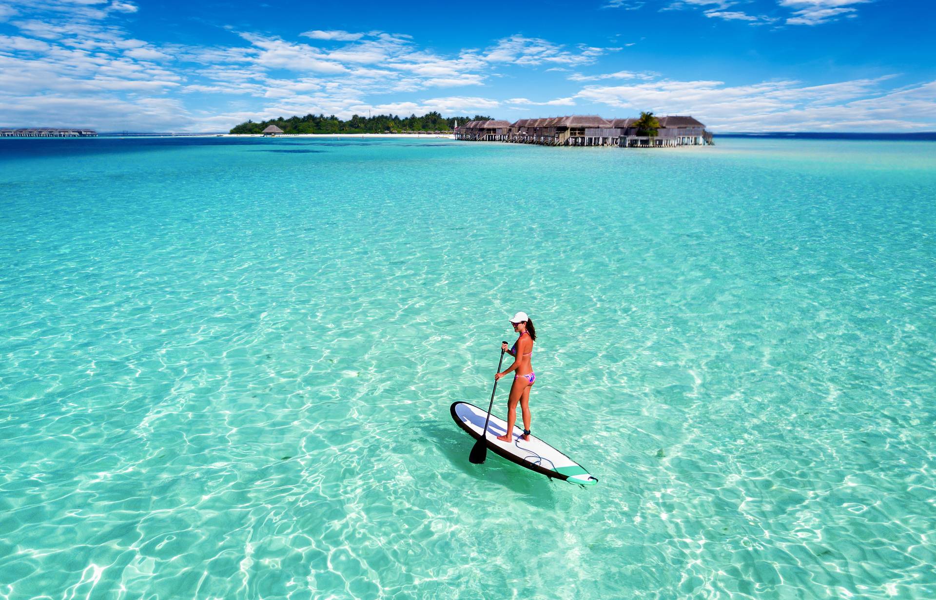 A woman paddleboards on crystal-clear turquoise waters with overwater bungalows in the distance.