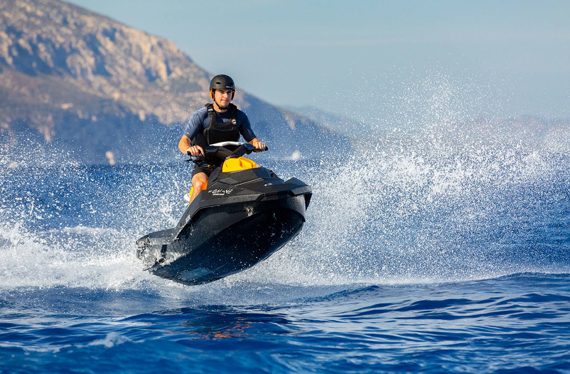 A man in a helmet and life vest expertly rides a jet ski through the choppy blue ocean, kicking up a dramatic spray of water.