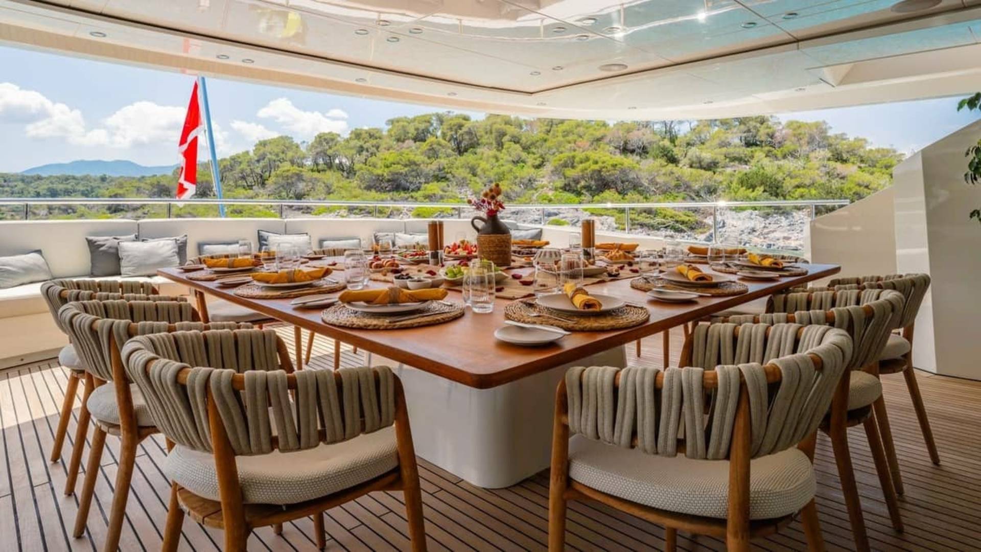 A beautifully set dining table on a yacht deck awaits guests with a stunning backdrop of lush green hills.