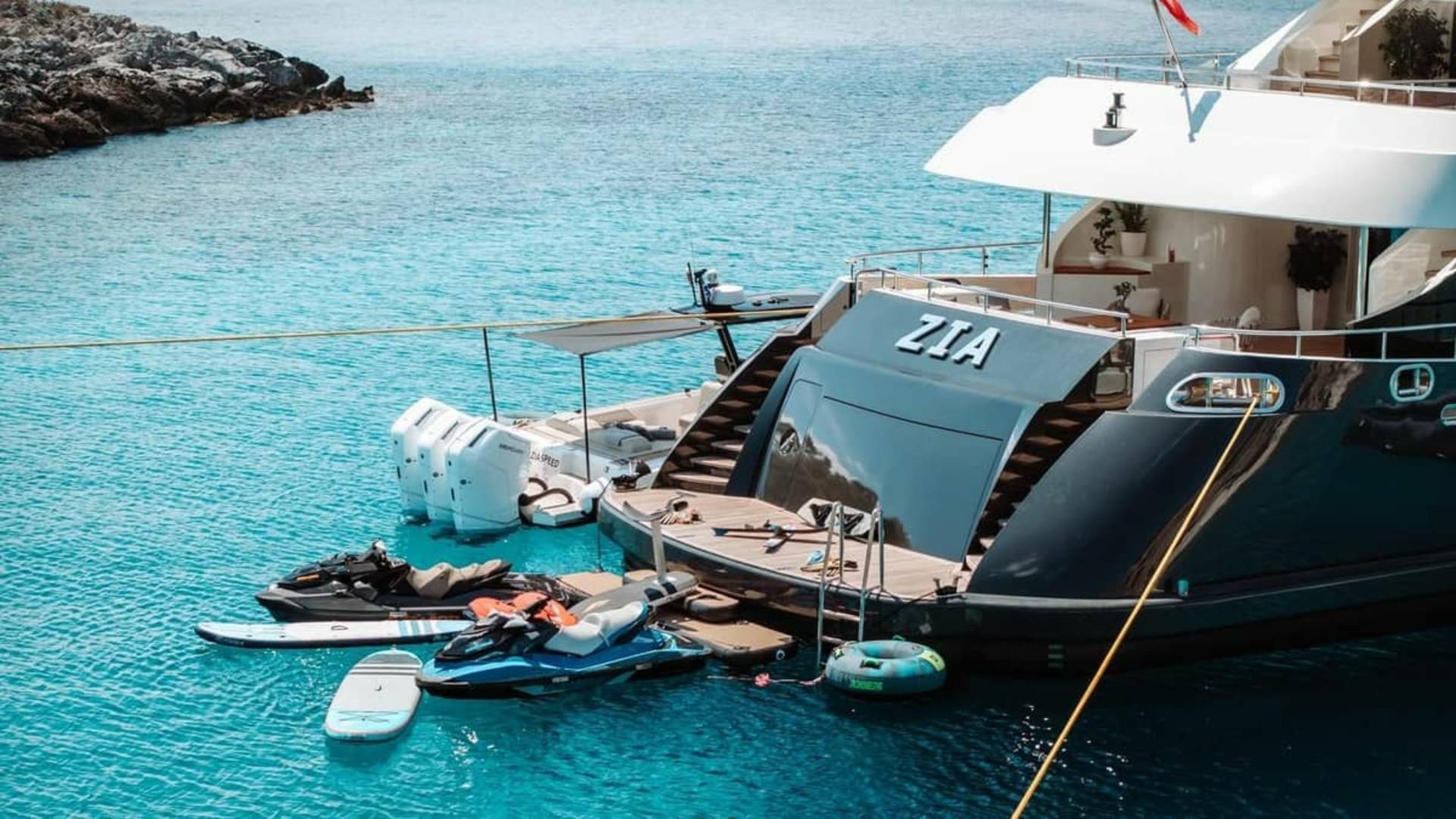 A luxurious yacht named Zia is anchored in crystal-clear turquoise waters, surrounded by jet skis and paddleboards.