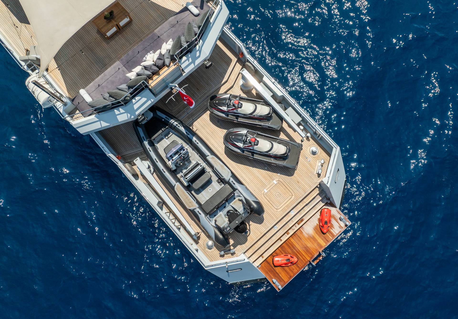 An aerial view captures a luxurious yacht adorned with jet skis and inflatable boats, floating on the sparkling blue ocean.