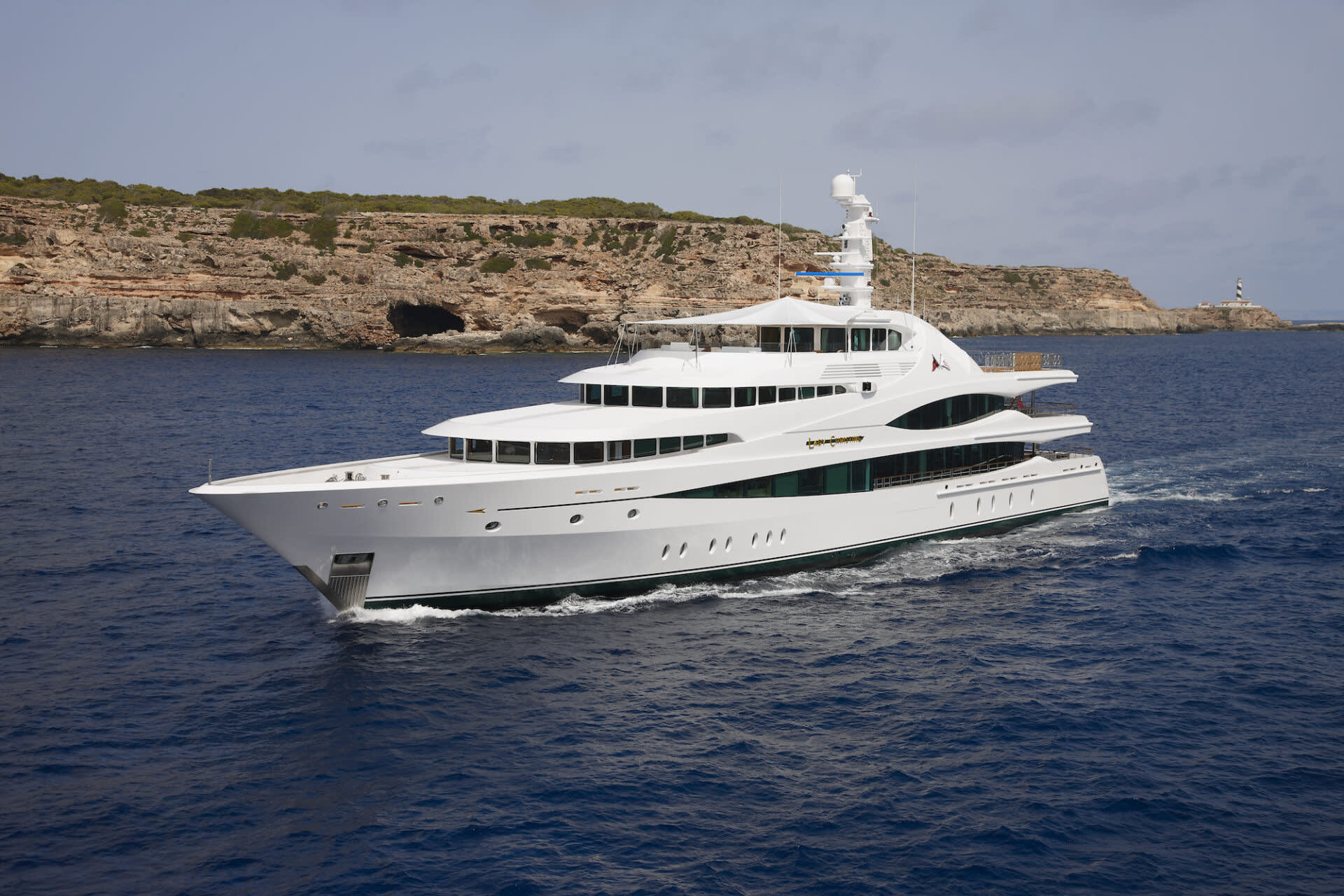 A luxurious white yacht glides through the deep blue sea near a rocky, vegetated coastline.