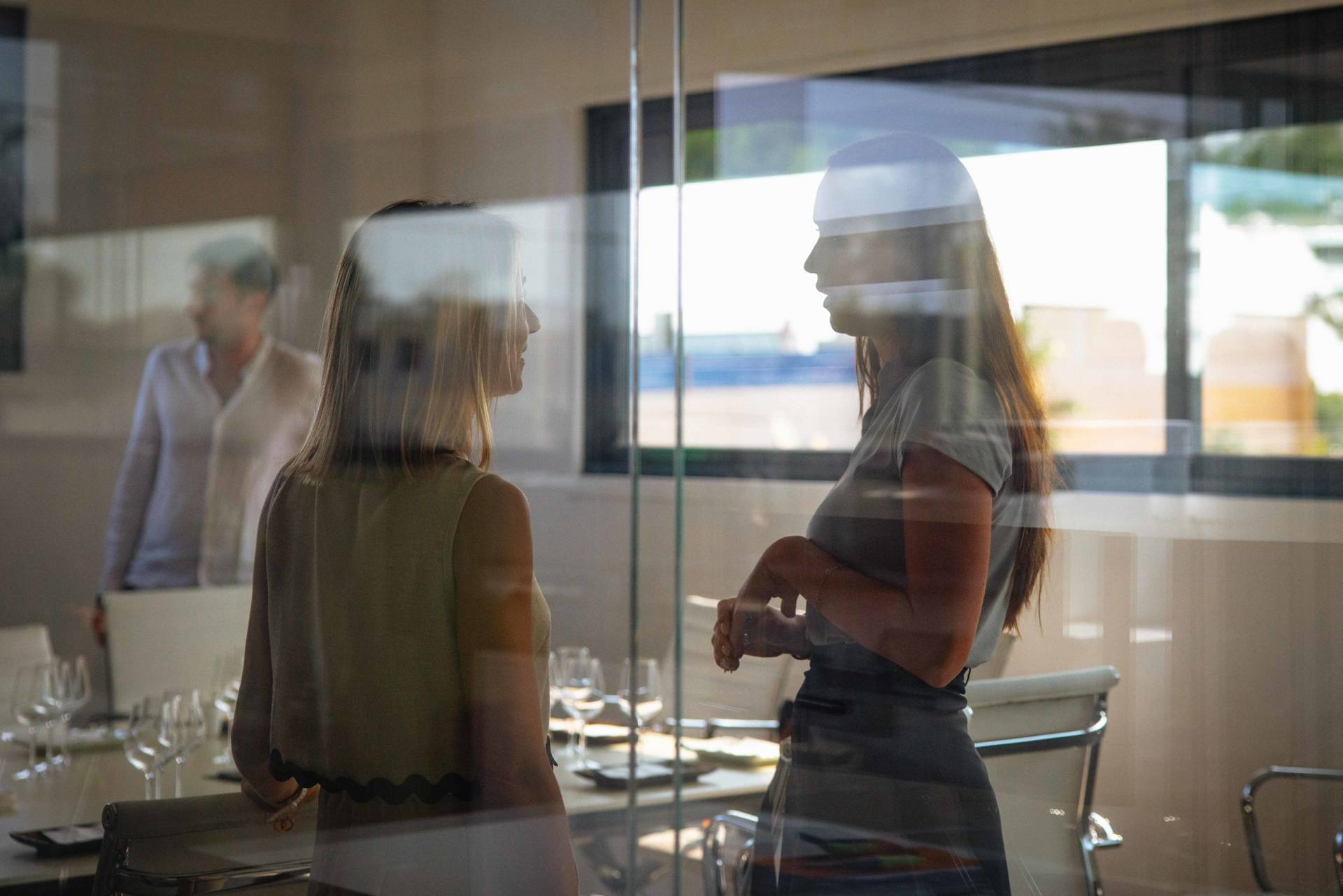 The image shows two people, a woman in a yellow sweater and a woman in a black top, standing in a room with a glass wall or window, with a blurred figure visible in the background.