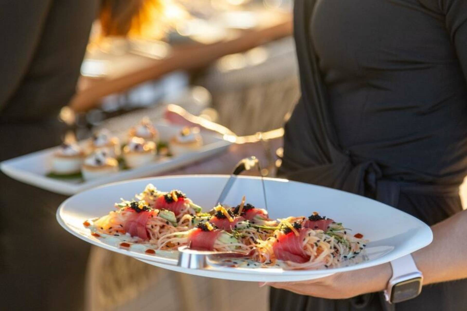 A server presents a plate of delicate tuna rolls topped with caviar and avocado, with a tray of appetizers in the background.