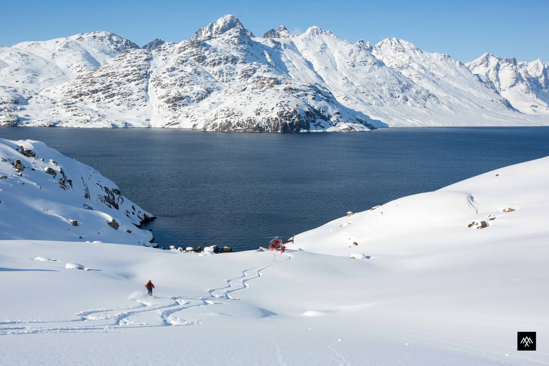 A lone skier carves fresh tracks down a snow-covered slope towards a deep blue fjord, with majestic, snow-capped mountains in the background.