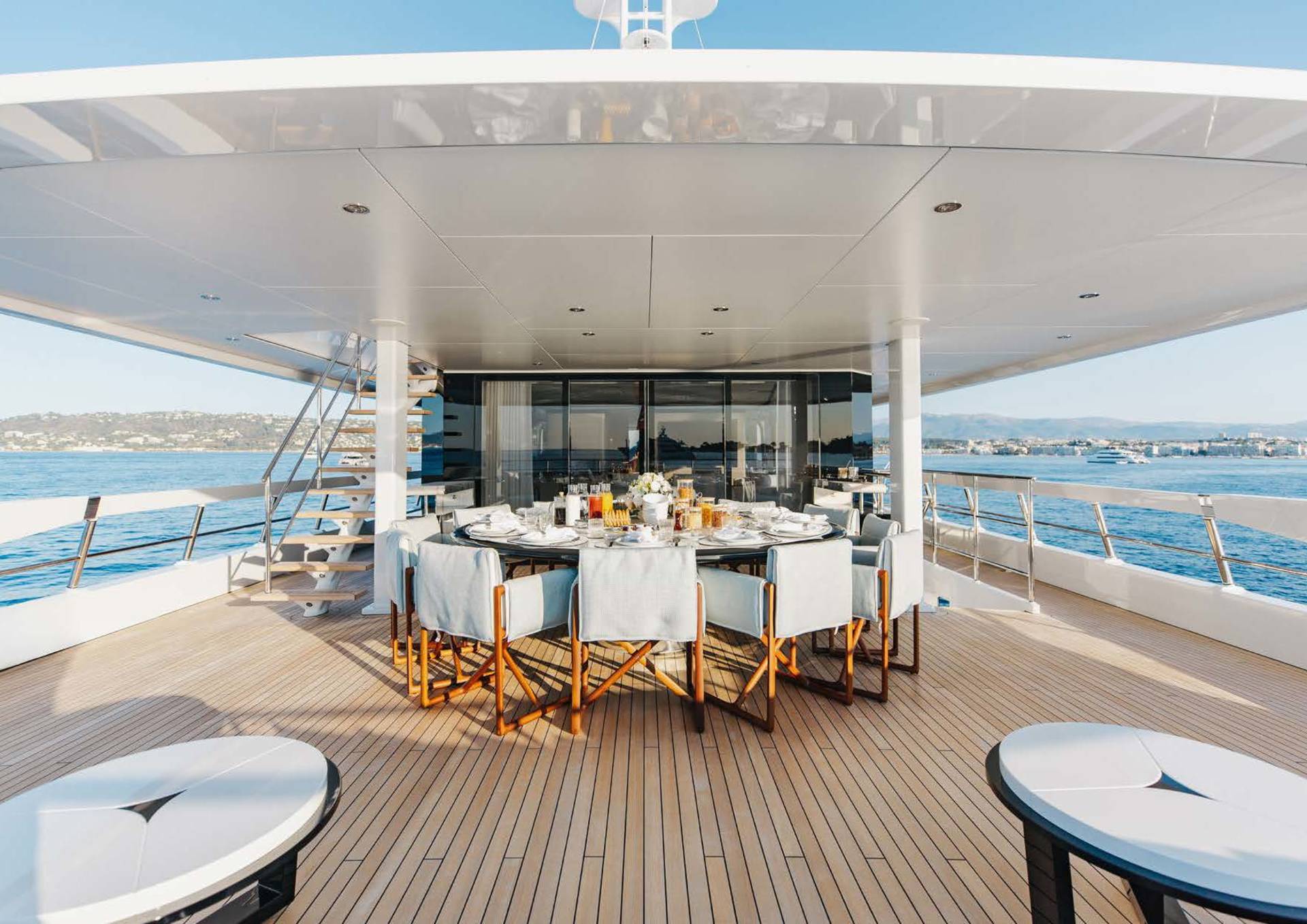 A lavishly set dining table awaits guests on the sun-drenched deck of a luxury yacht overlooking a picturesque coastline.