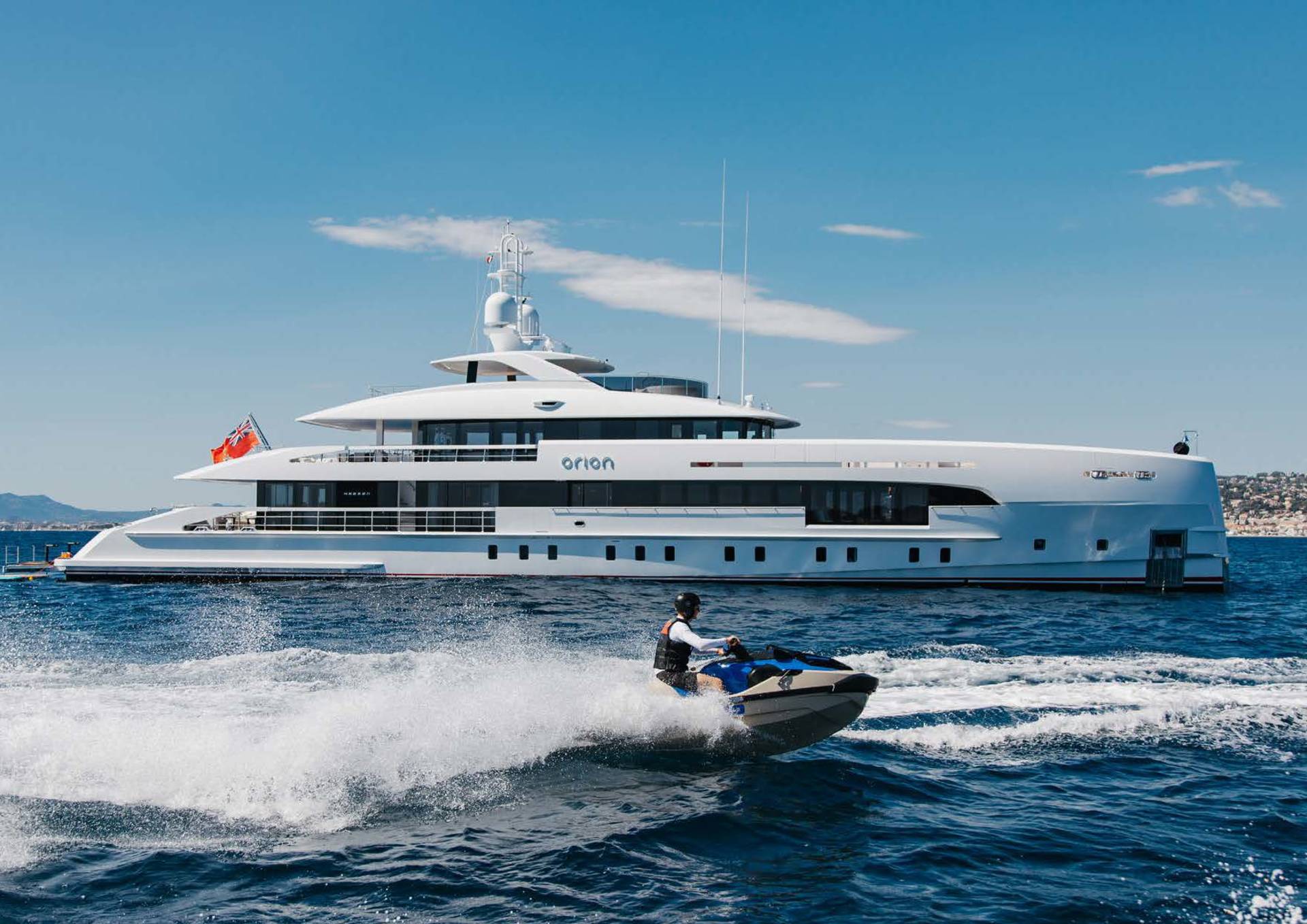 A person on a jet ski creates a spray of water in front of a large white yacht named Orion under a bright blue sky.
