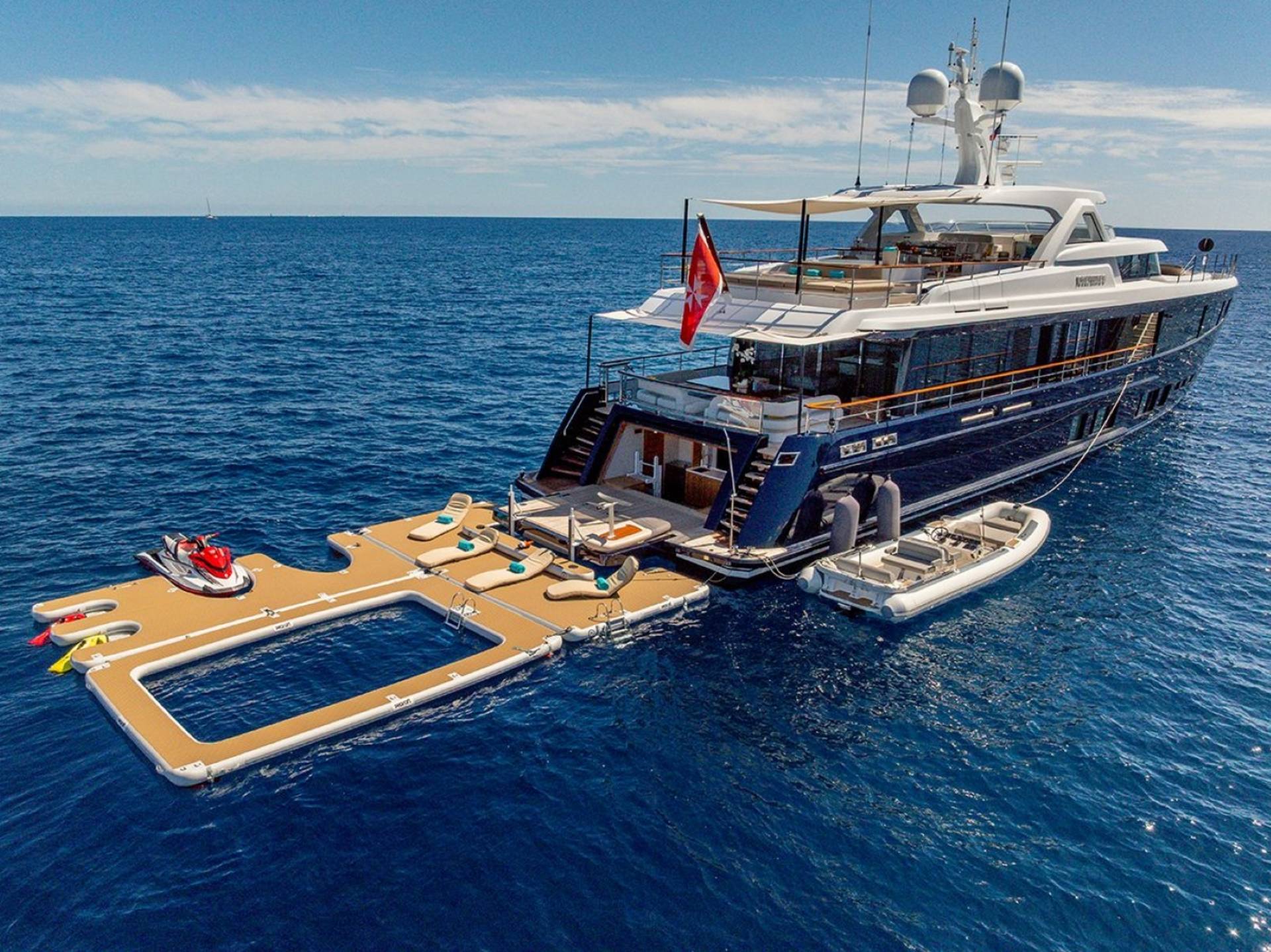 A luxurious yacht anchors in the deep blue sea, featuring a floating deck with a pool, loungers, and a jet ski.