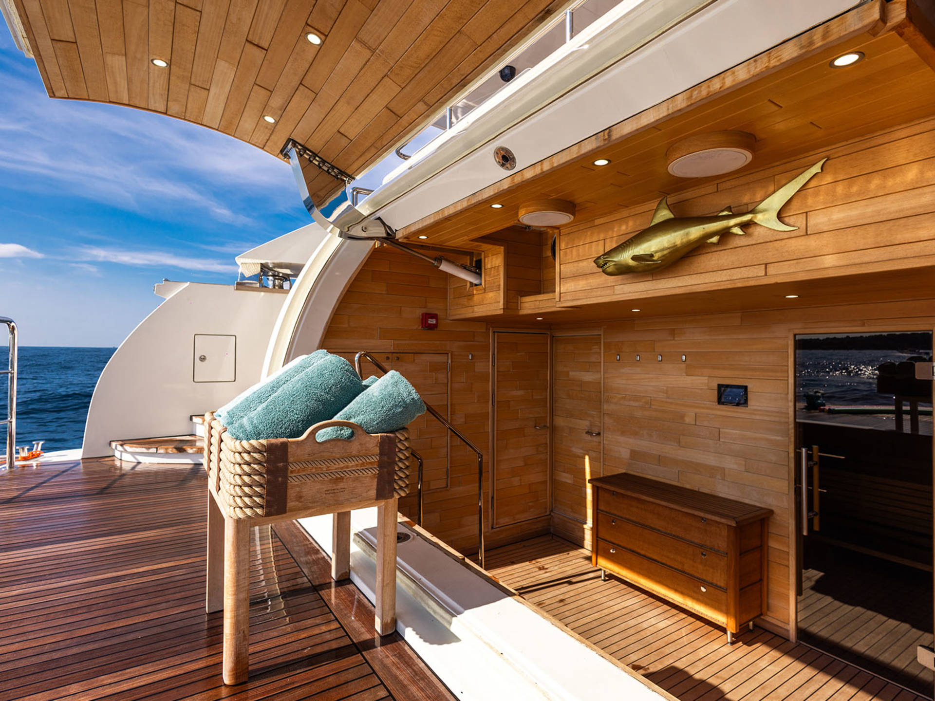A luxurious yacht deck features teal towels in a wooden basket, a golden shark sculpture, and a polished wooden chest.