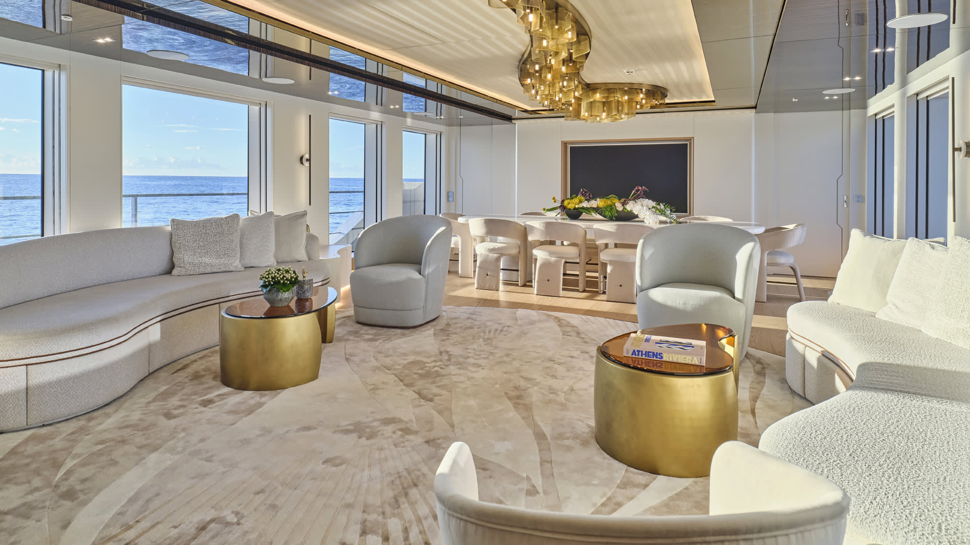 A luxurious and spacious living room with large windows overlooking the ocean, featuring plush white furniture, gold accents, and a grand chandelier.