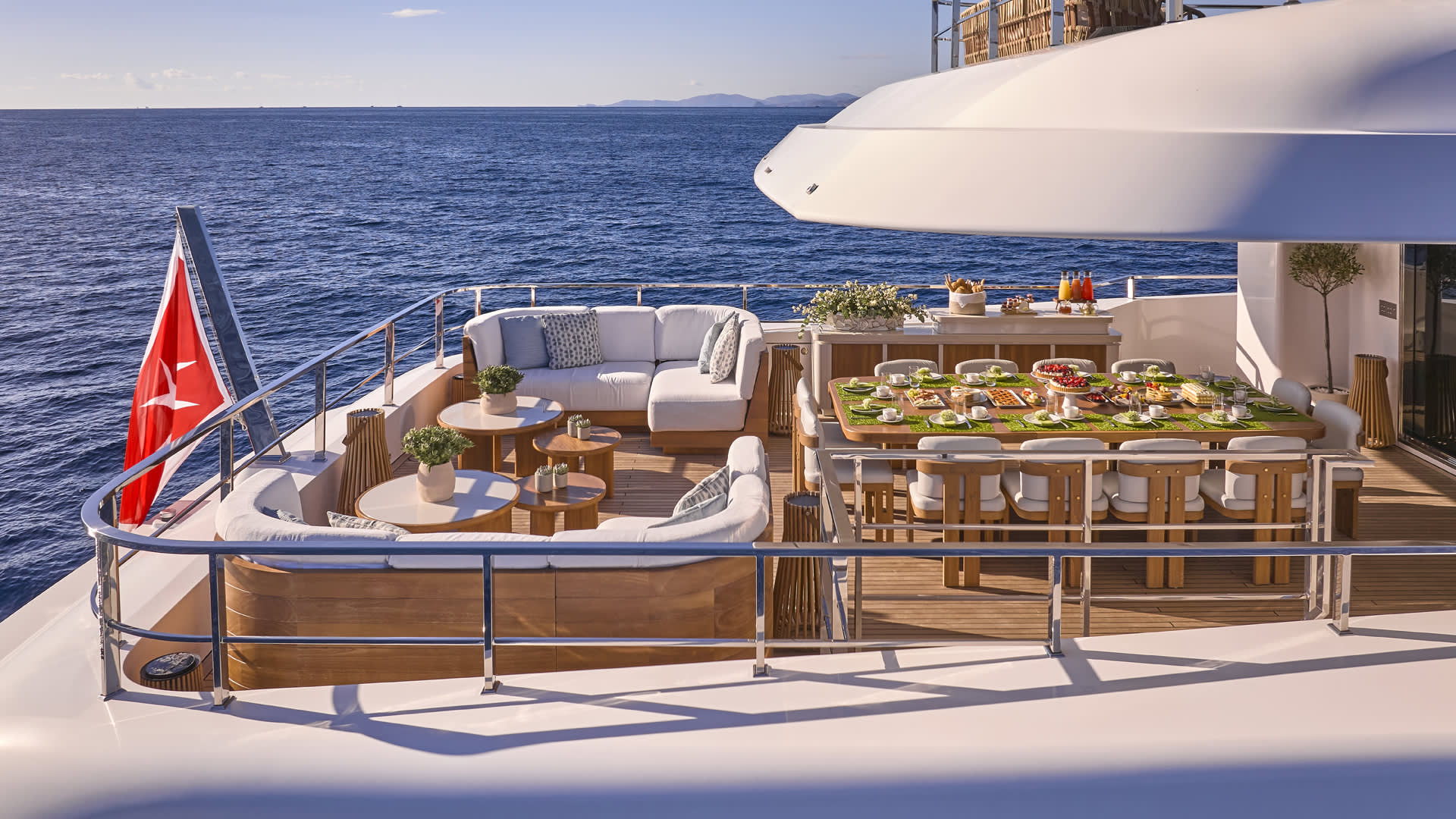 A luxurious yacht with a spacious outdoor deck, featuring comfortable seating, a dining table, and a scenic backdrop of the open sea and distant mountains.