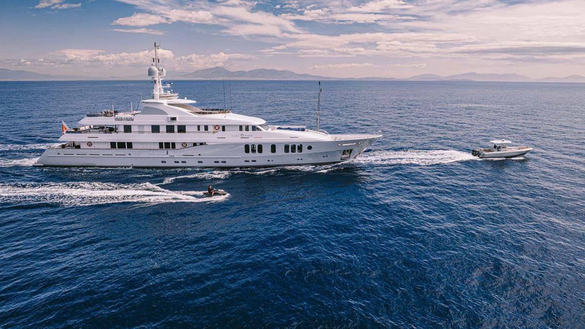 A luxurious superyacht cruises through the deep blue sea, trailed by a smaller boat and a jet ski.