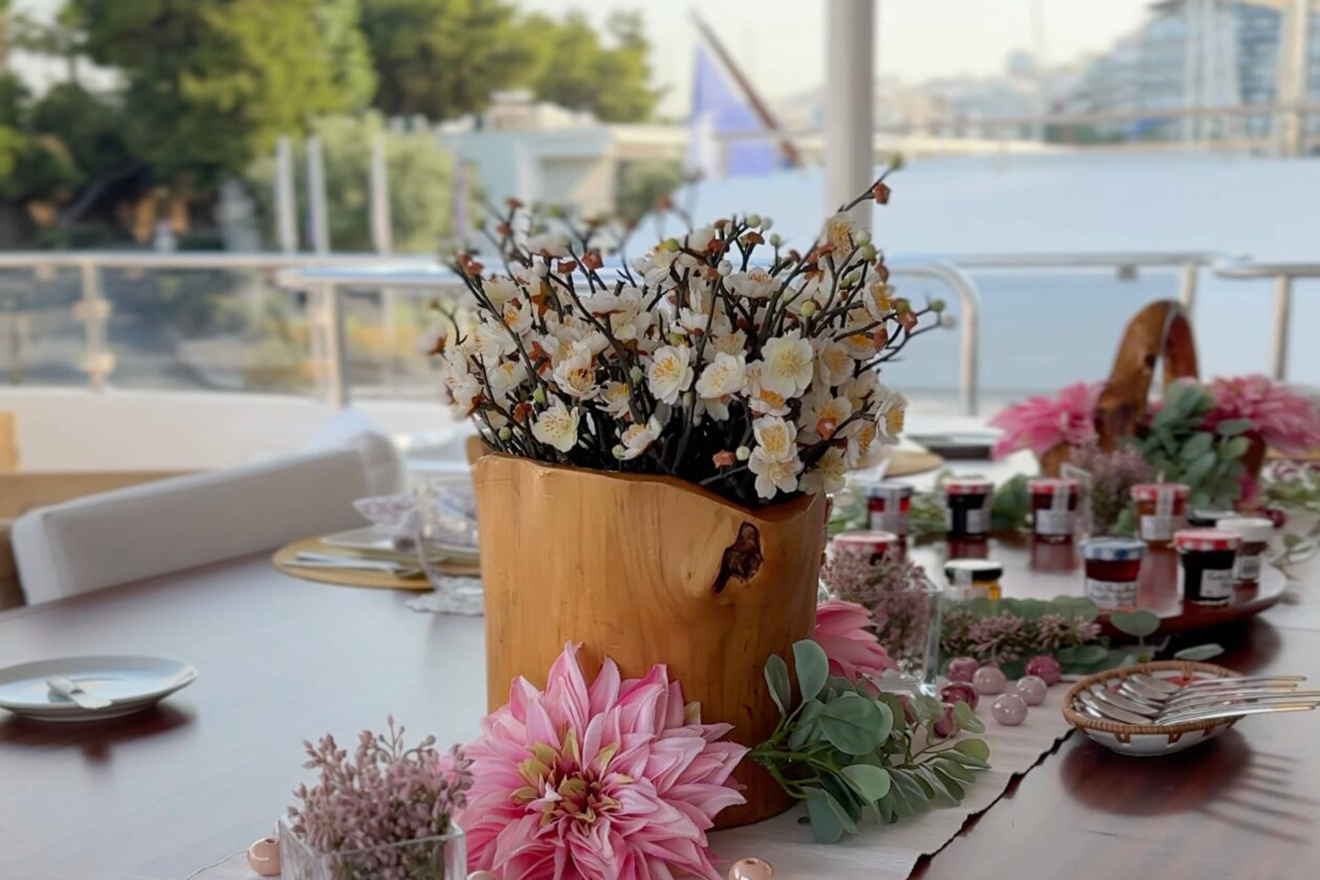 A beautifully set table with floral arrangements and jars of jam awaits guests on a sunny deck overlooking a harbor.