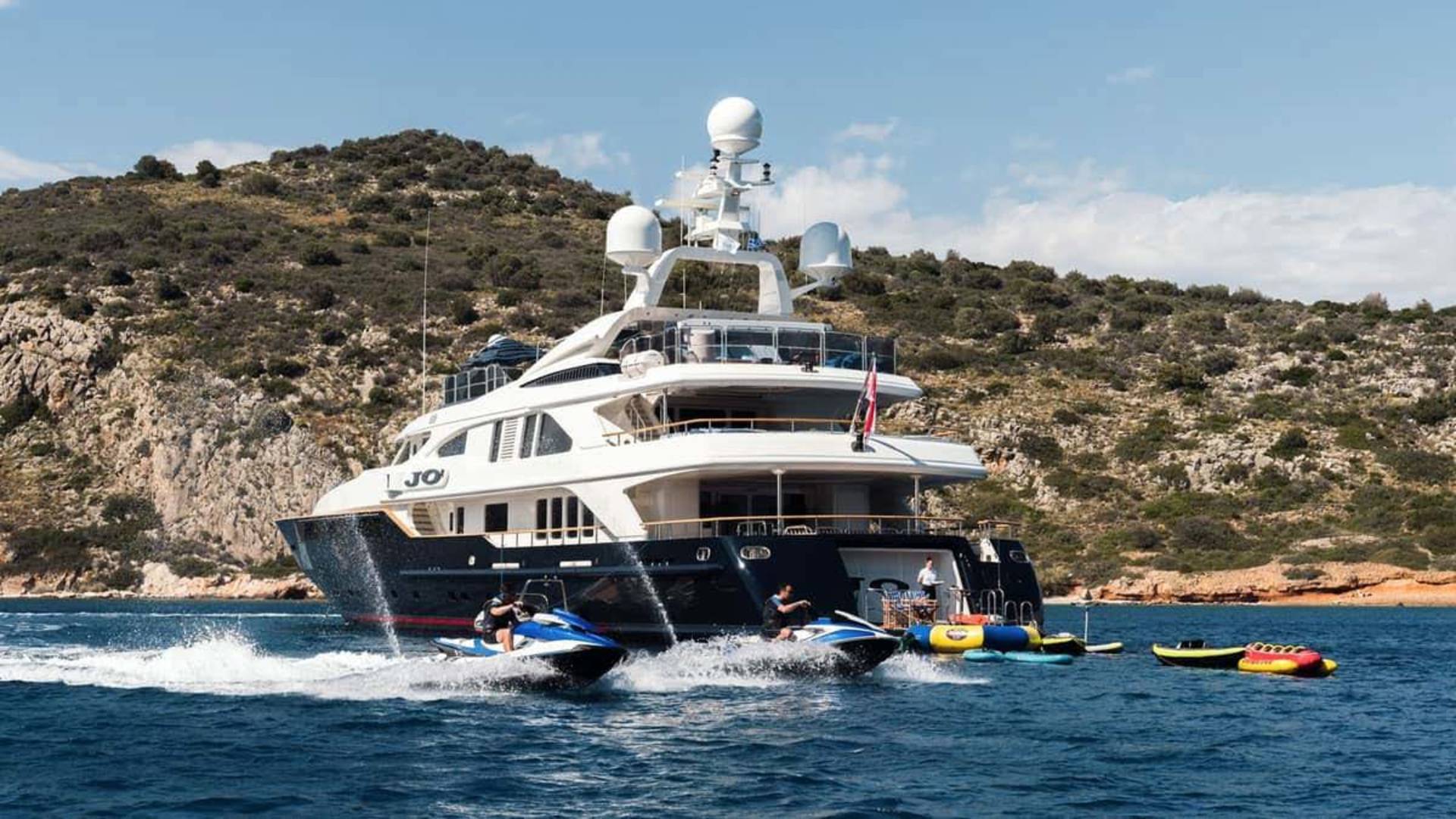 Jet skis and inflatable toys create a lively scene around a luxurious yacht anchored near a rocky, shrub-covered island.
