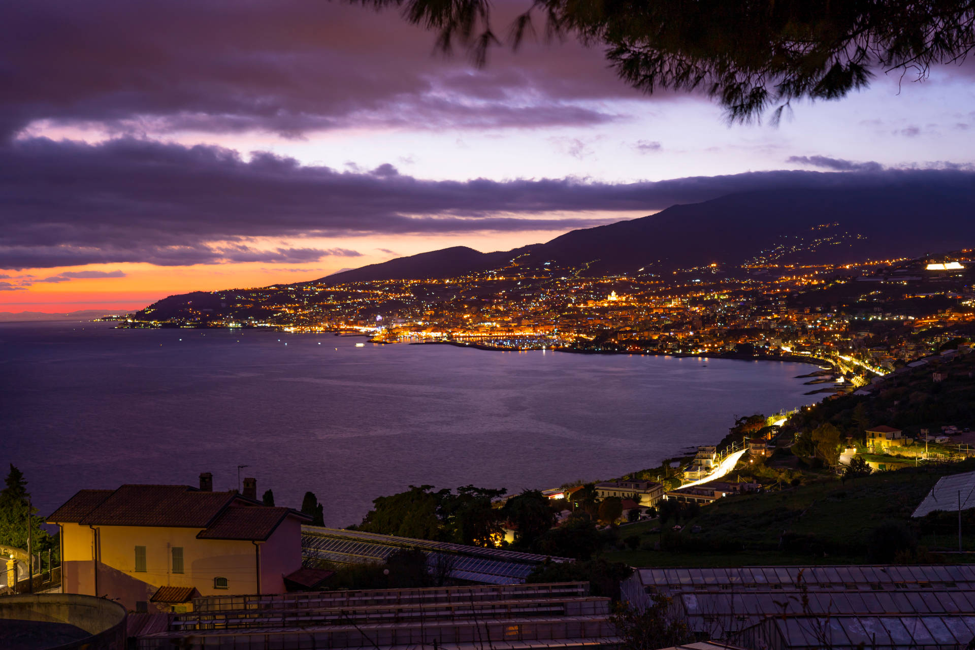 A coastal city glows with warm lights under a dramatic twilight sky as the sun dips below the horizon.