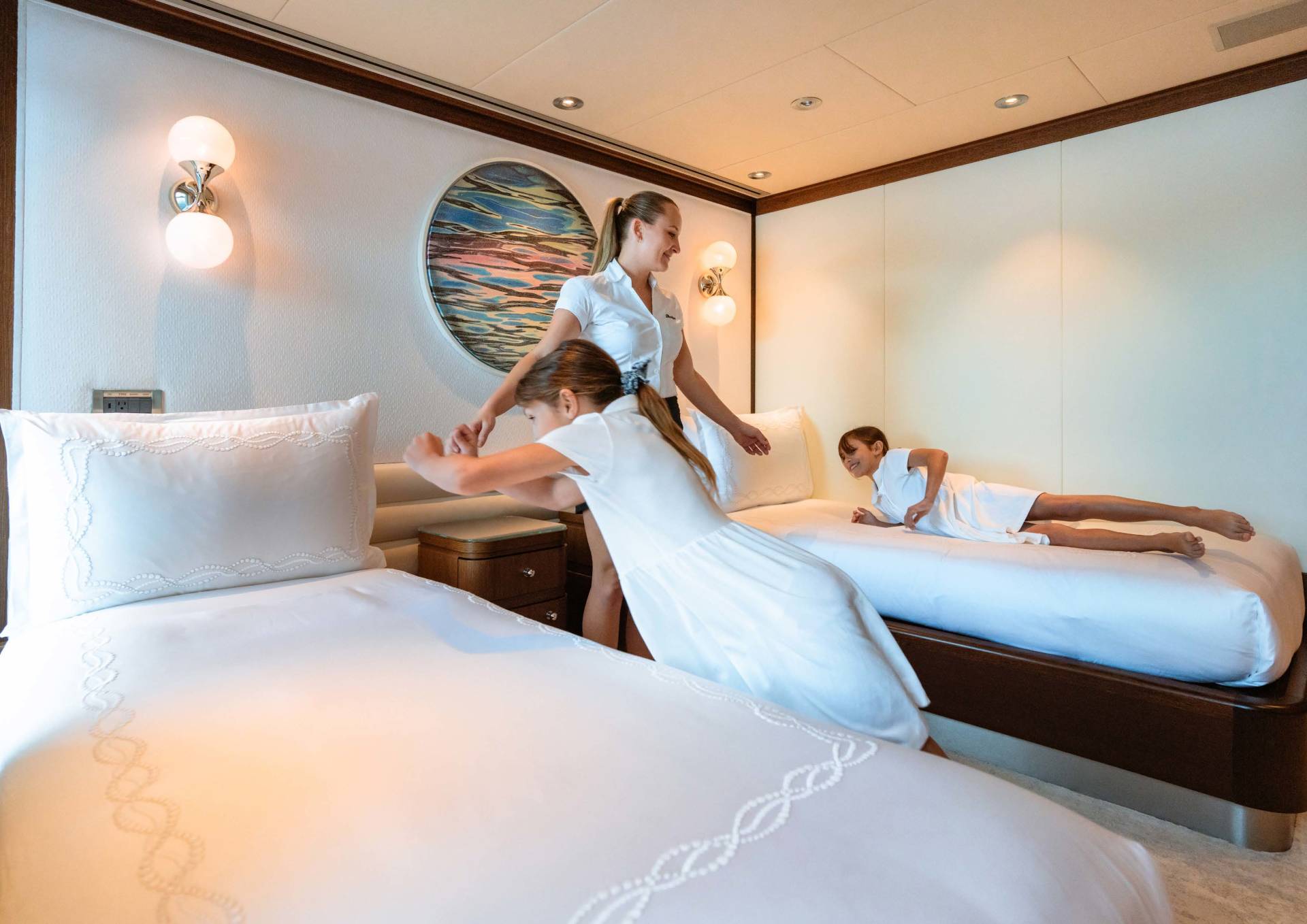 A woman in uniform playfully interacts with two children in a luxurious cabin with twin beds.