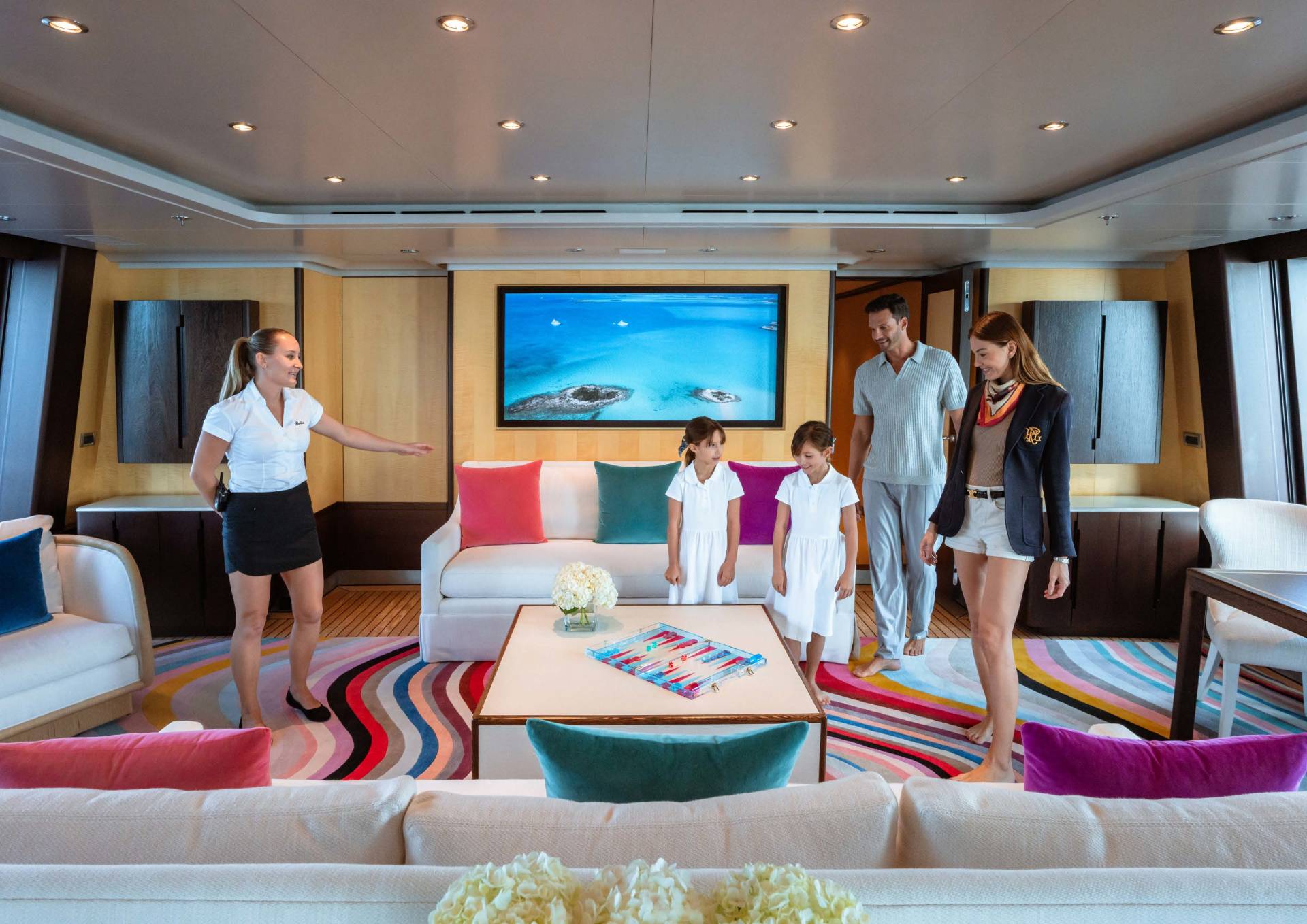 A family enjoys a luxurious yacht interior with a vibrant rug and a large screen displaying a tropical seascape.