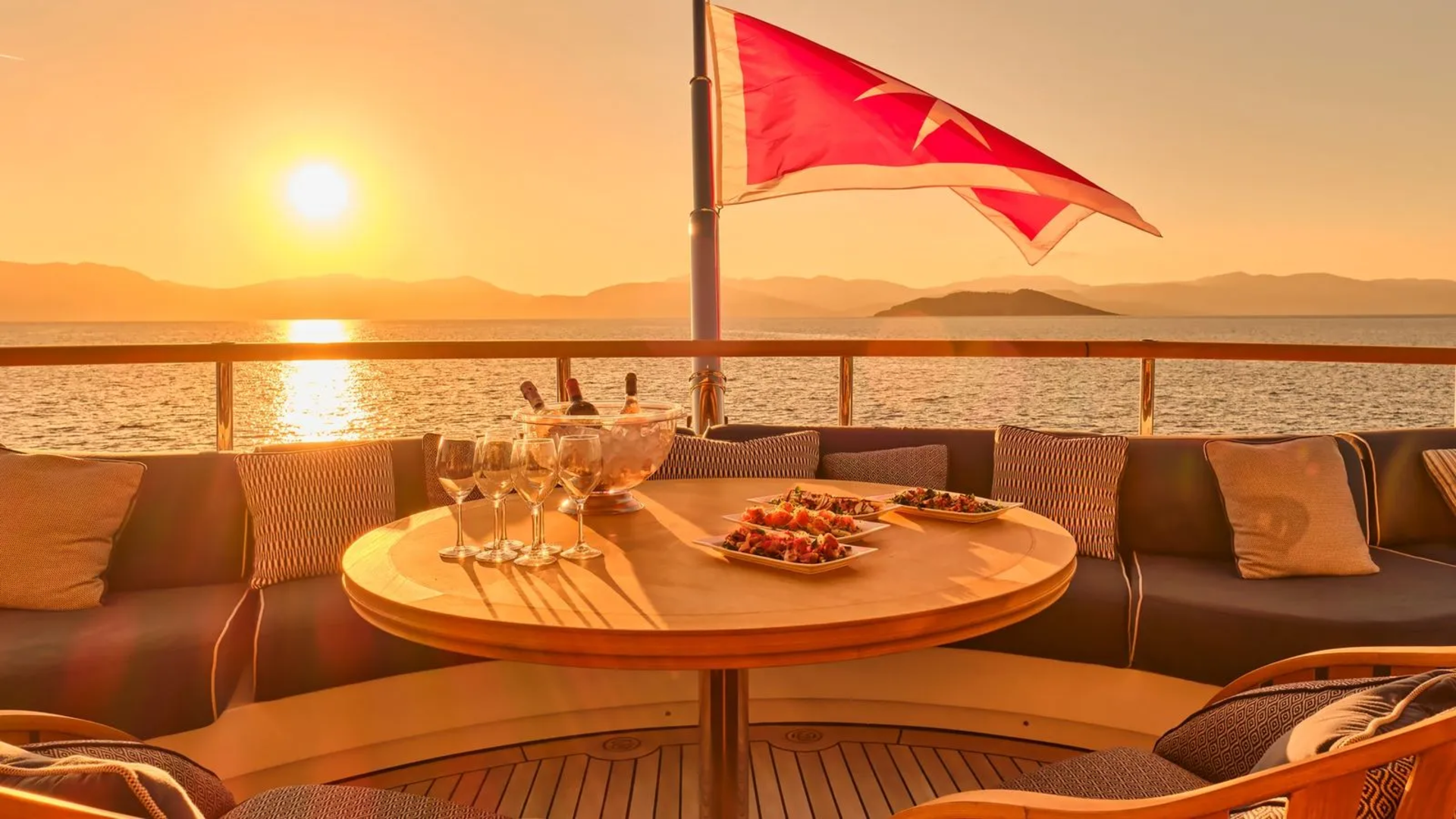A Turkish flag waves over a yacht's deck as the sun sets, casting a warm glow on a table set for a luxurious meal.