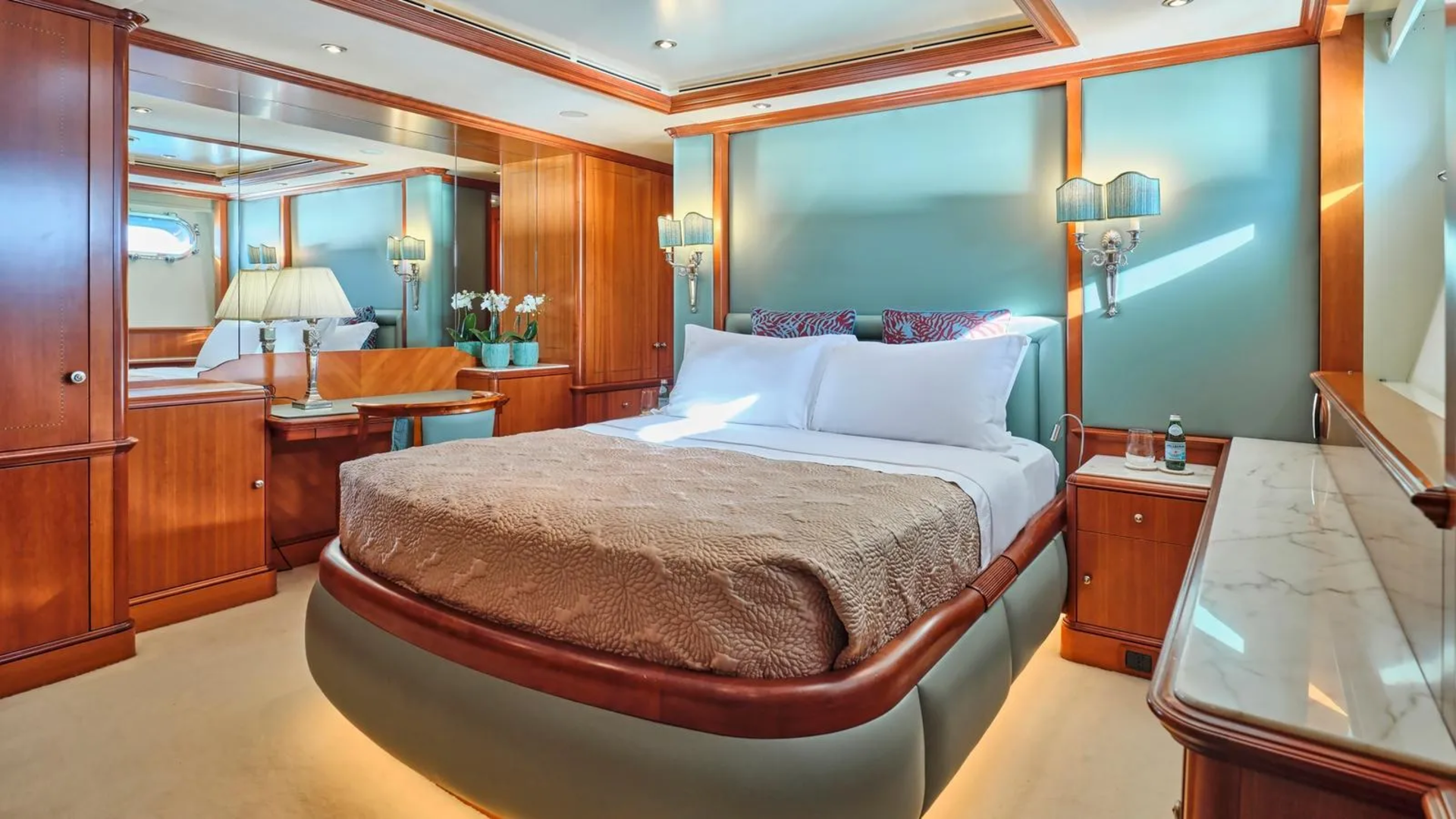 Luxurious yacht cabin features a plush bed, elegant wood finishes, and serene teal walls bathed in natural light.