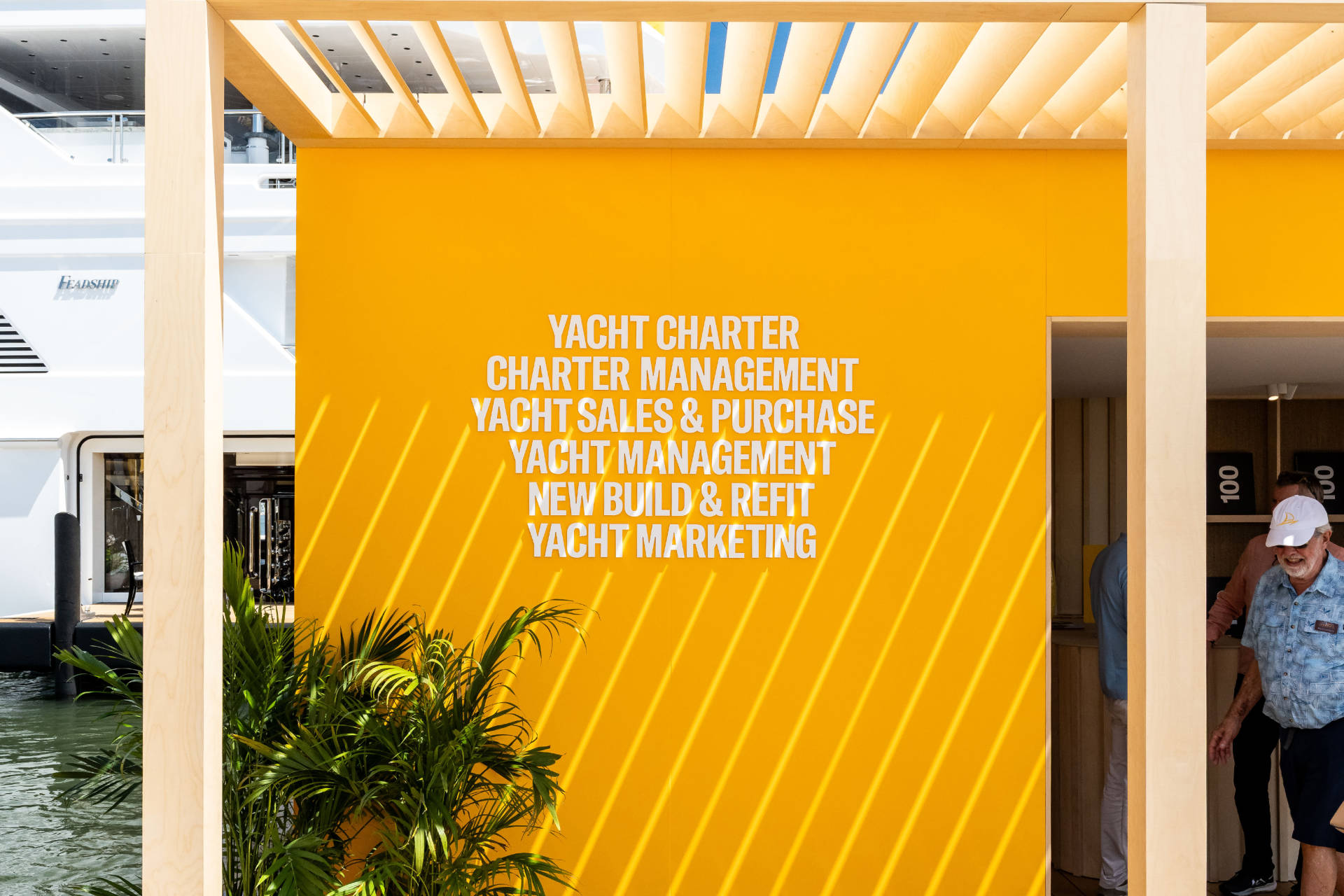 A bright yellow wall with a list of yacht services in white text, partially obscured by a wooden trellis and tropical plants.