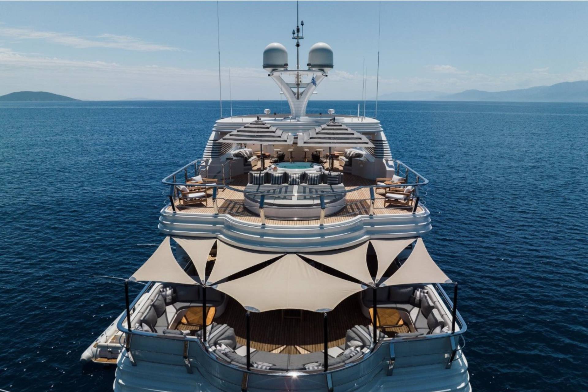A luxurious yacht with multiple decks and shaded seating areas cruises on calm blue waters under a clear sky.