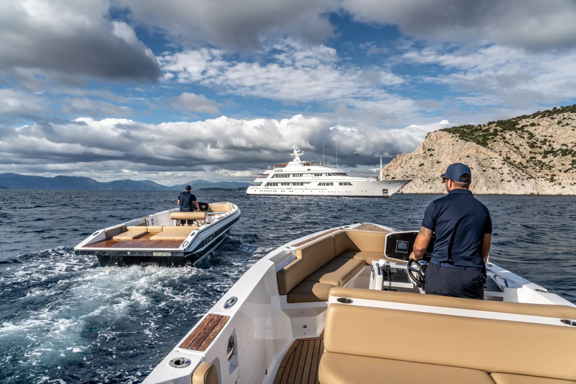 A large white luxury yacht is seen in the background, while in the foreground, two smaller boats are navigating the choppy waters against a backdrop of cloudy skies and mountainous terrain.