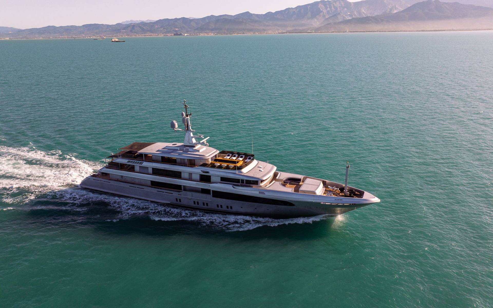 A large, luxurious yacht is speeding across a serene, turquoise-colored body of water, with a mountainous landscape visible in the background.