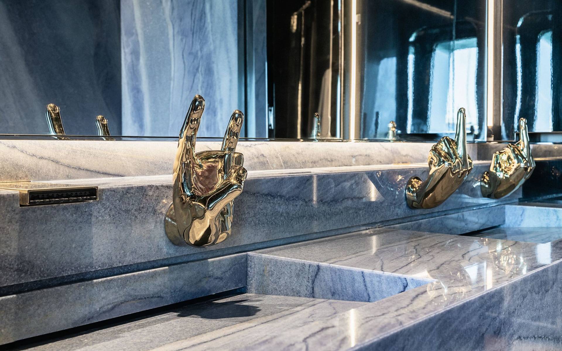A row of golden faucet handles on a marble countertop, with a mirrored background reflecting the scene.