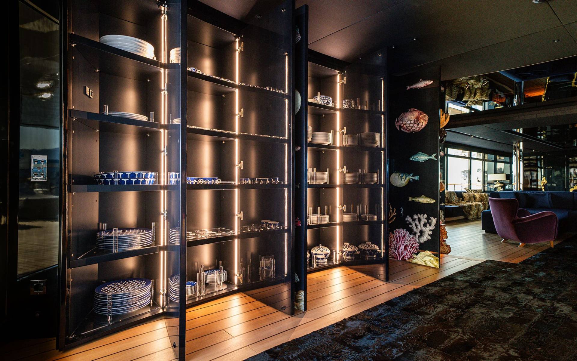 A dimly lit interior space with a large glass-enclosed shelving unit displaying various glassware and decorative items, set against a backdrop of a cozy lounge area with plush seating.