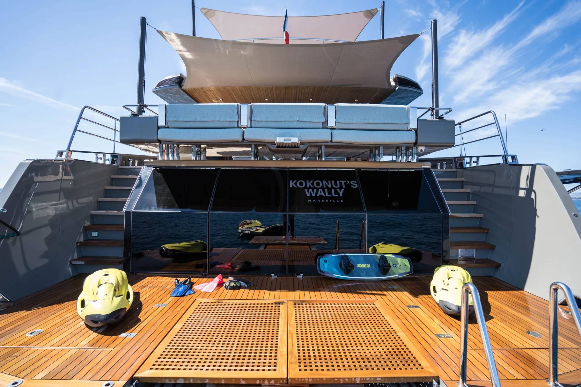 Two yellow jet skis and water sports equipment rest on the wooden deck of a yacht, with the yacht's name "KOKONUT'S WALLY" visible on a dark panel in the background.