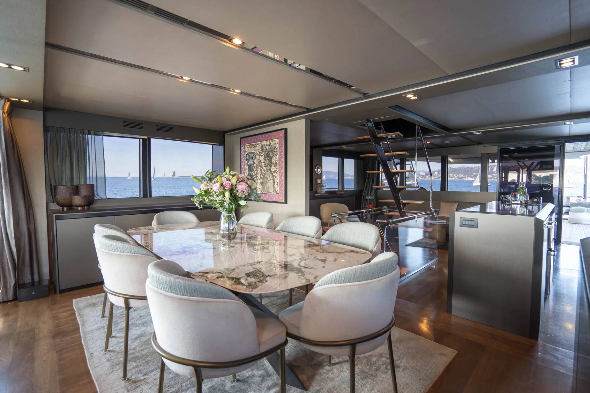 A luxurious dining room with a marble-topped table and plush chairs in the foreground, and a staircase leading to an upper level with a view of sailboats on the ocean in the background.